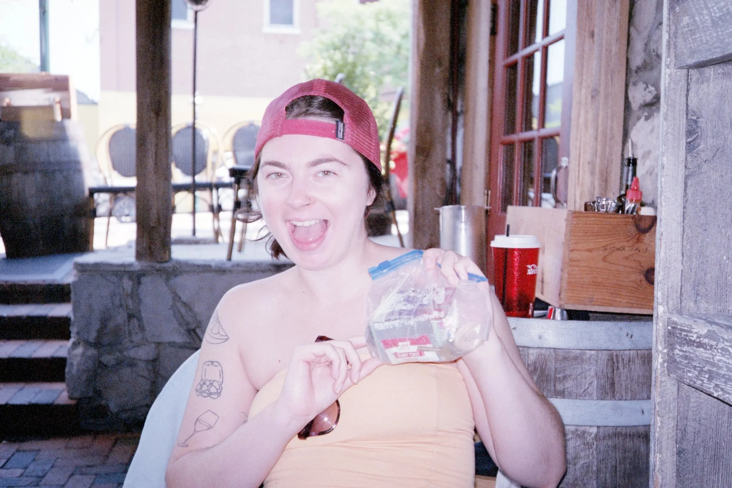 A woman with short dark hair wearing a red baseball cap and a strapless orange top, smiling and holding her purse, which is a plastic bag, at an outdoor restaurant.