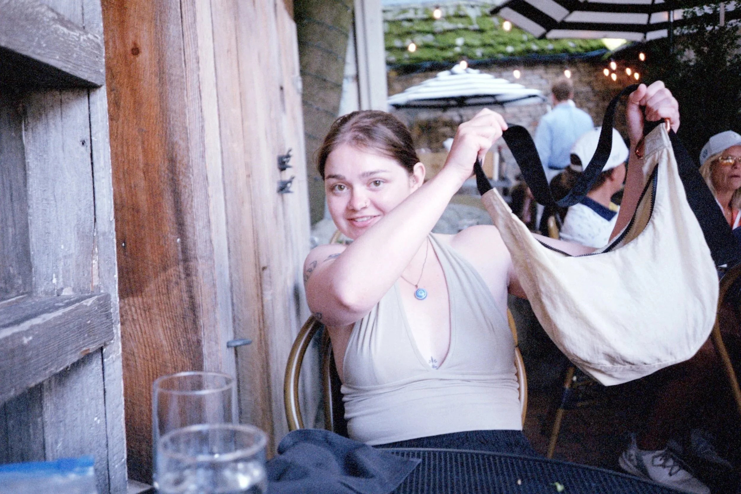 A woman with brown hair and a necklace holding a cream Baggu bag at an outdoor restaurant.