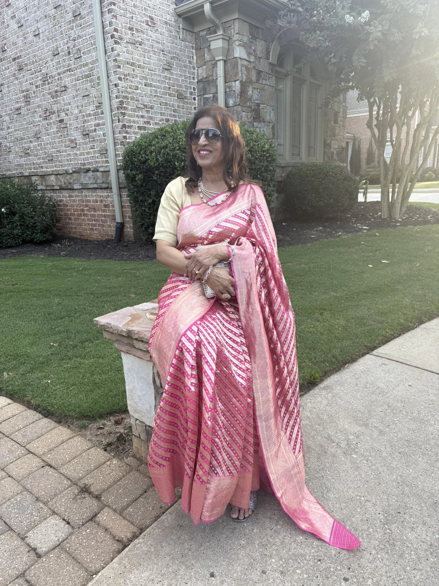 Woman in a pink and gold traditional Indian saree with sunglasses sitting on a stone bench outside a brick house.