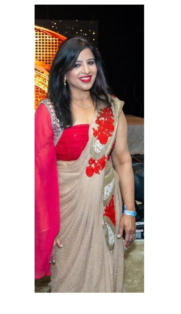 A woman wearing a beige saree with red embroidery and a red blouse, smiling at the camera, standing indoors. She has black hair, earrings, and a wristwatch.