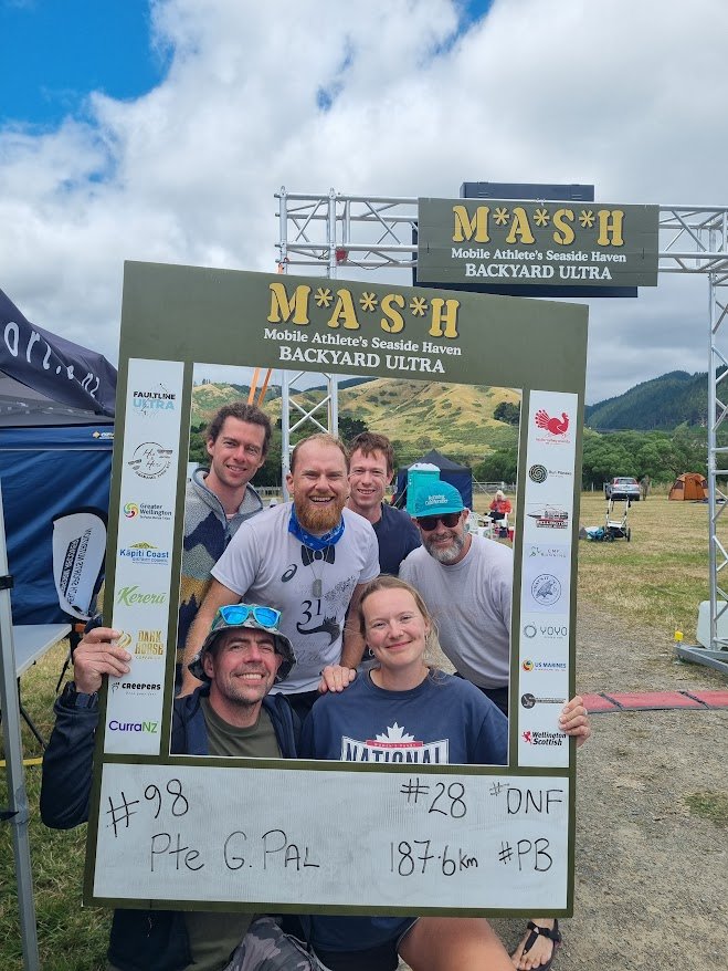 Gordon posing with a group of five people posing behind a large decorative frame at the M*A*S*H Backyard Ultra race event, outdoors with tents, cars, and mountains in the background, after completing 28 laps or 187km.