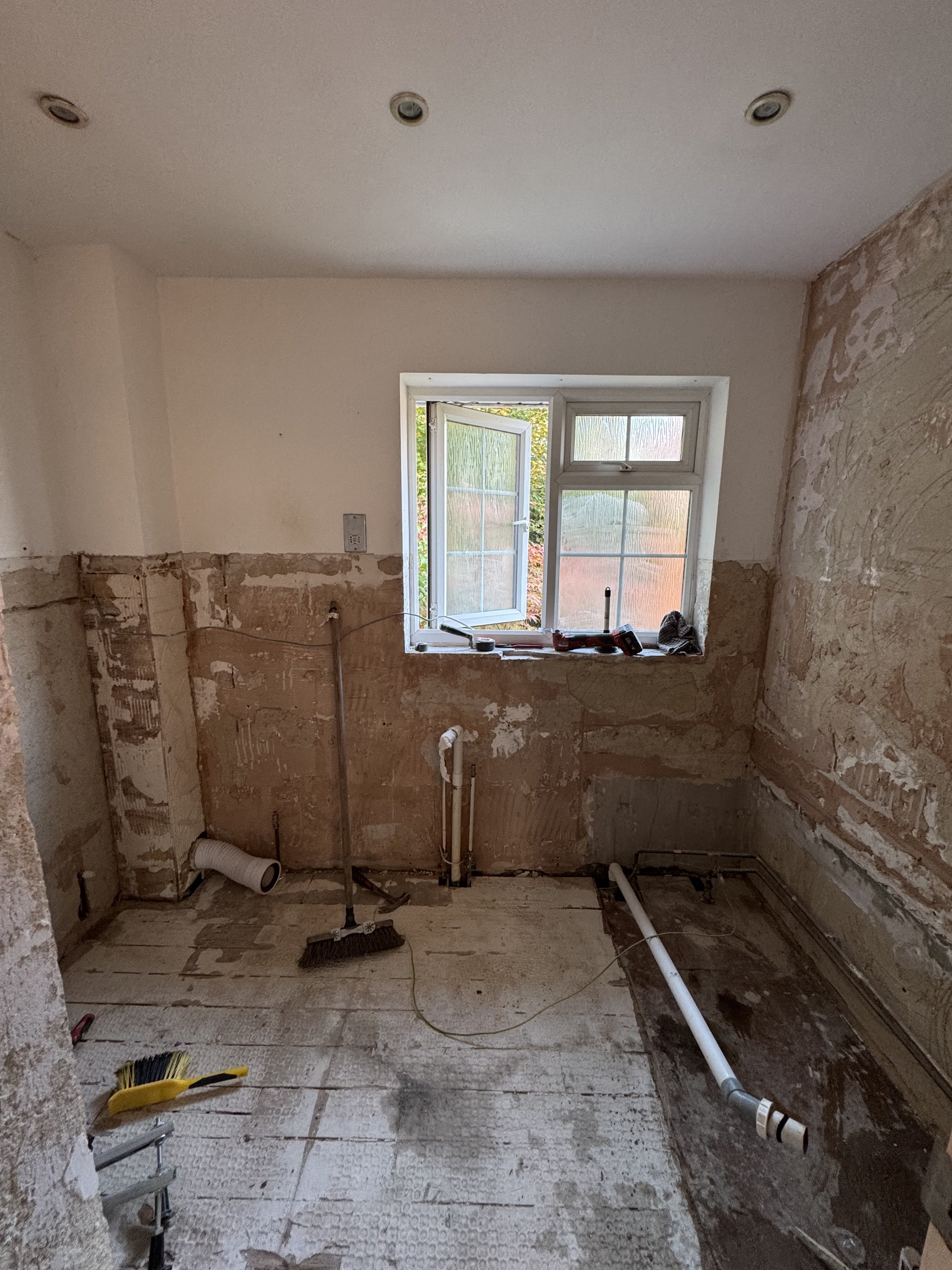 Room under renovation with exposed walls, pipes, and plumbing fixtures, window open, construction tools on windowsill, and construction debris on the floor.