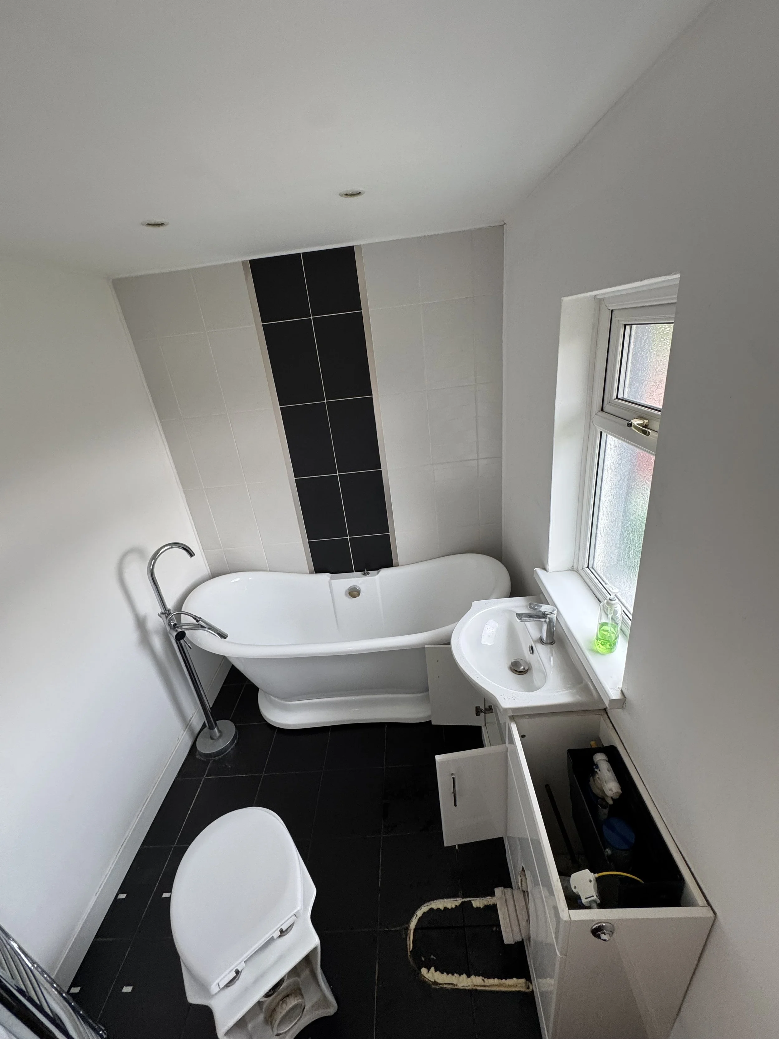 A small bathroom with white walls and black tile floors. It has a white bathtub with a curved edge, a small white sink with a cabinet underneath, and a white toilet with the lid open. There is a small window on the right side with a bottle of green liquid soap on the windowsill. The bathroom appears to be under renovation, with a cabinet door open and some plumbing visible.