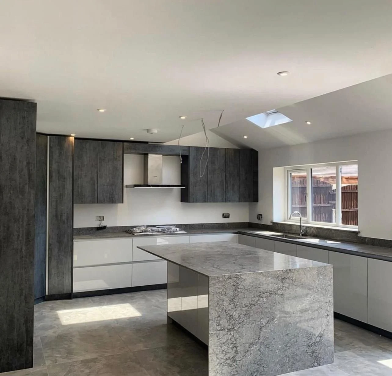Modern kitchen with grey and white cabinets, a large marble island, and a window above the sink showing a wooden fence outside.