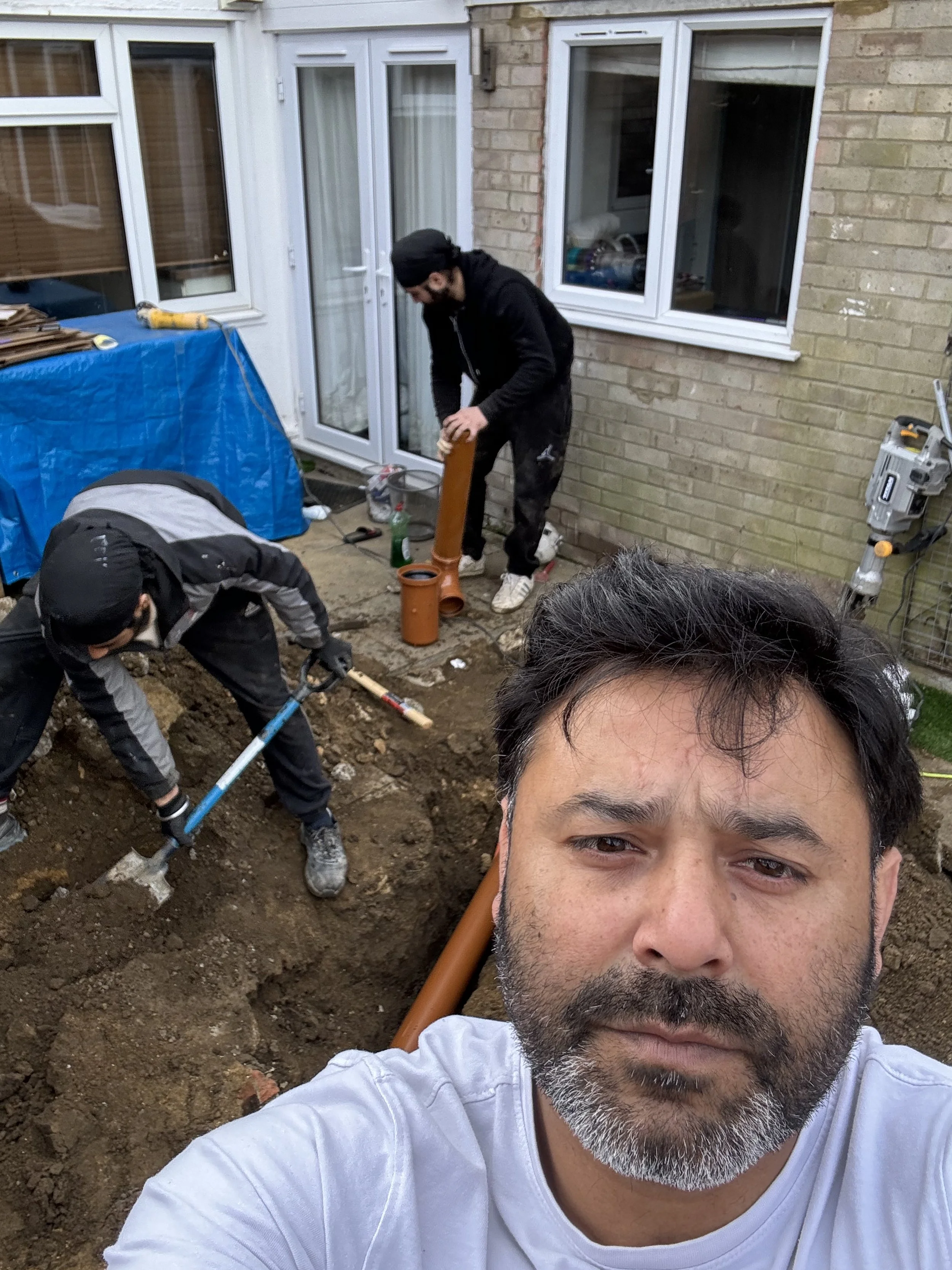 A man taking a selfie in a backyard under renovation, with two other workers digging and installing pipes in the dirt, and a house exterior with windows and a door in the background.