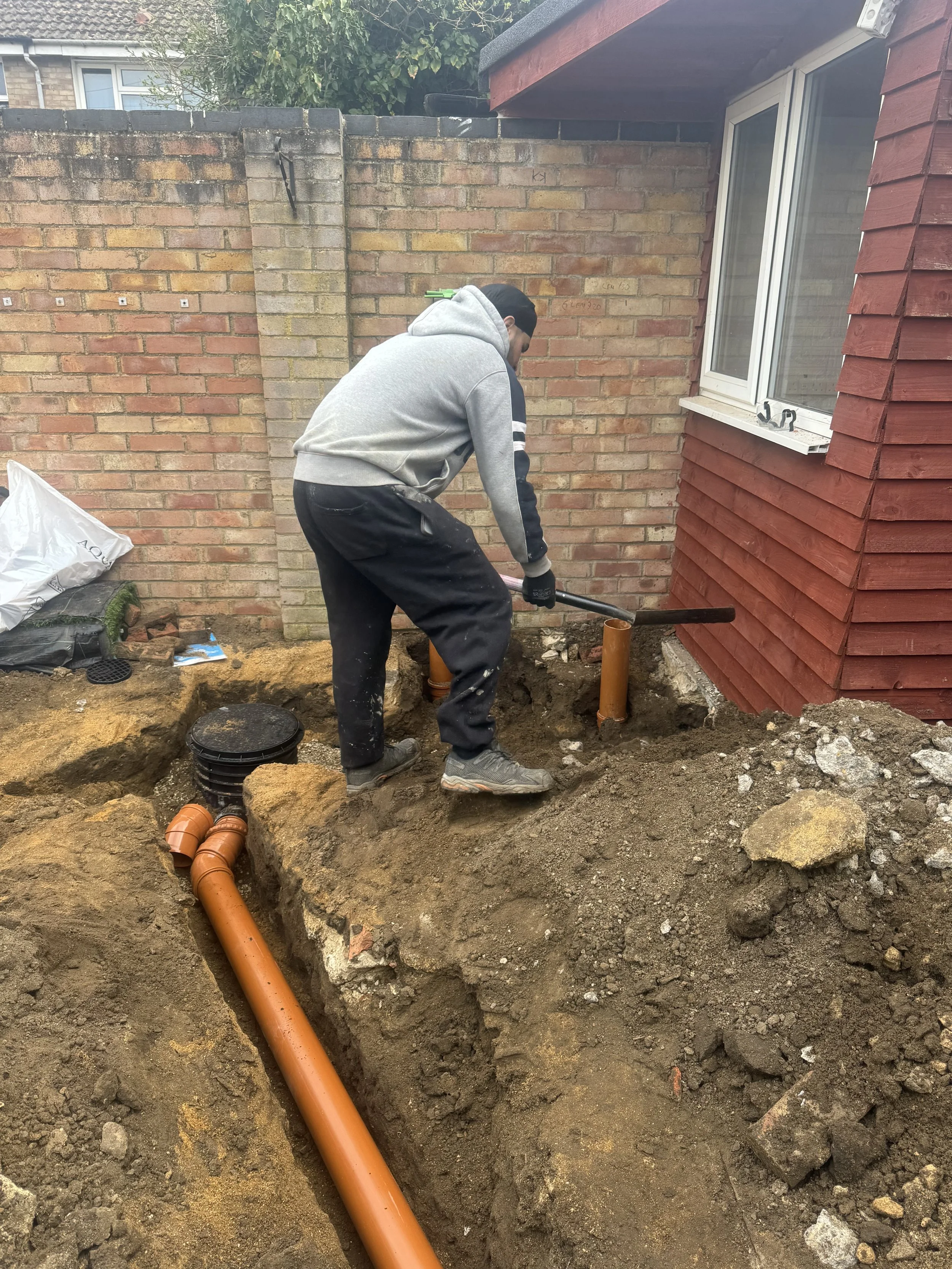 A man working on plumbing outside a house, digging a trench in the dirt near the house foundation. He is using a pickaxe, and there are orange pipes in the trench and lying nearby. The house has red wooden siding, and there is a brick wall behind him.
