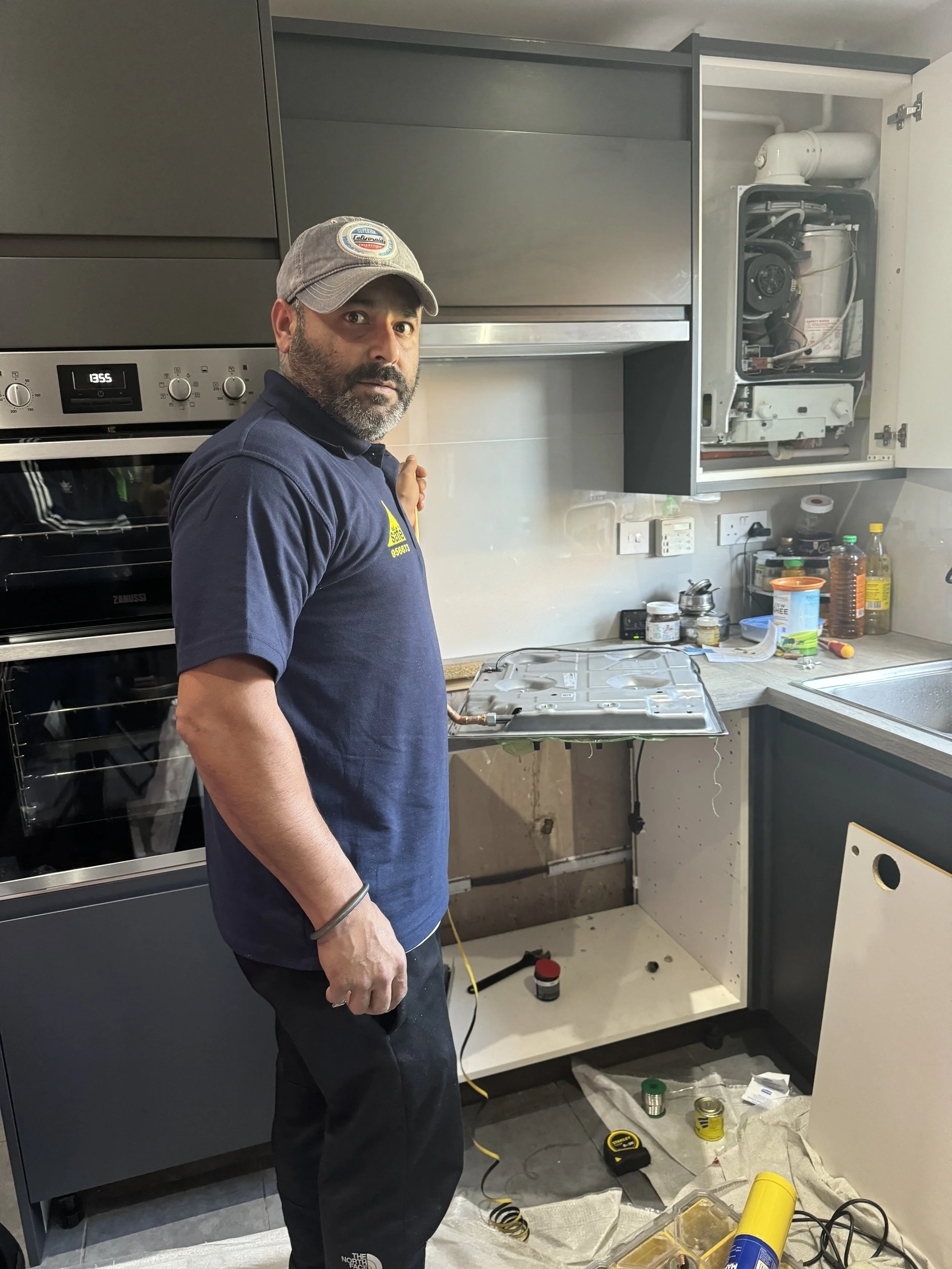 A man wearing a cap and navy polo shirt stands in a kitchen under repair, holding a metal panel. The kitchen has an oven, open cabinets with appliances, and tools and supplies scattered on the counter and floor.