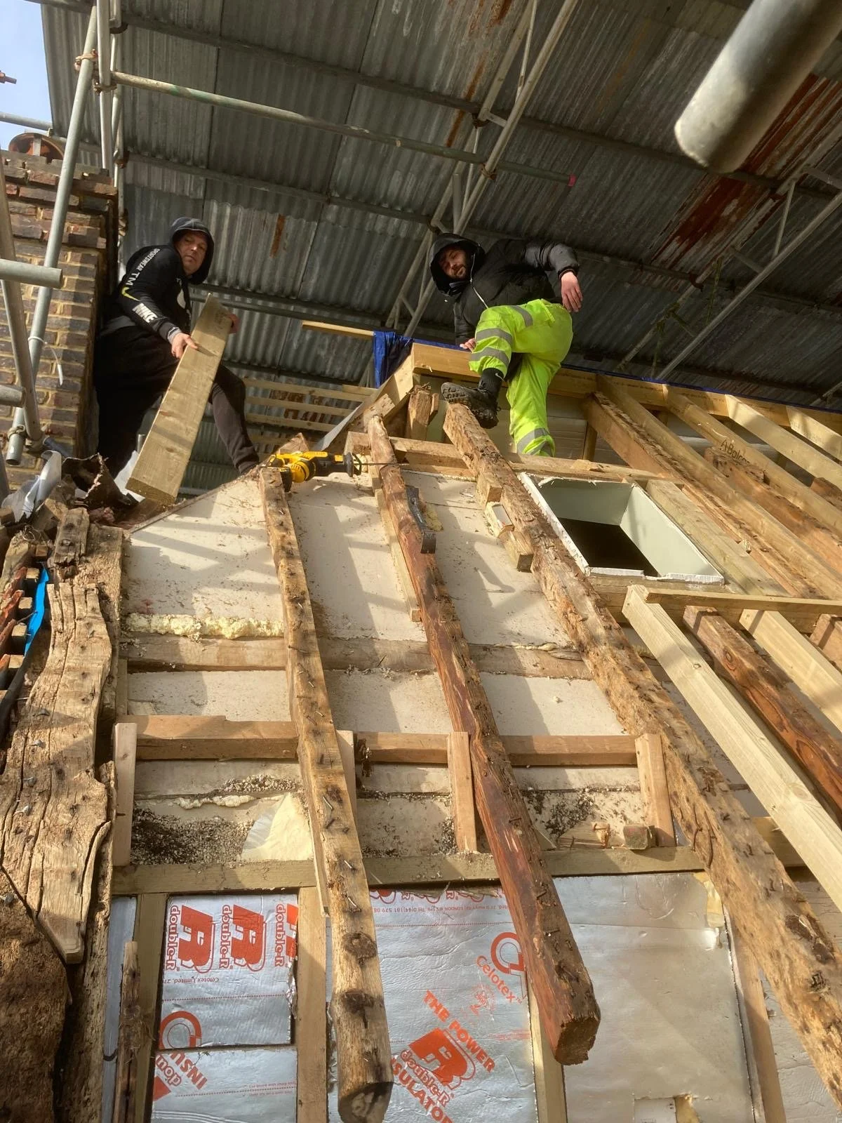 Three construction workers are working on a building frame inside a covered area. They are standing on the wooden framework of the wall, with construction tools and materials around them.