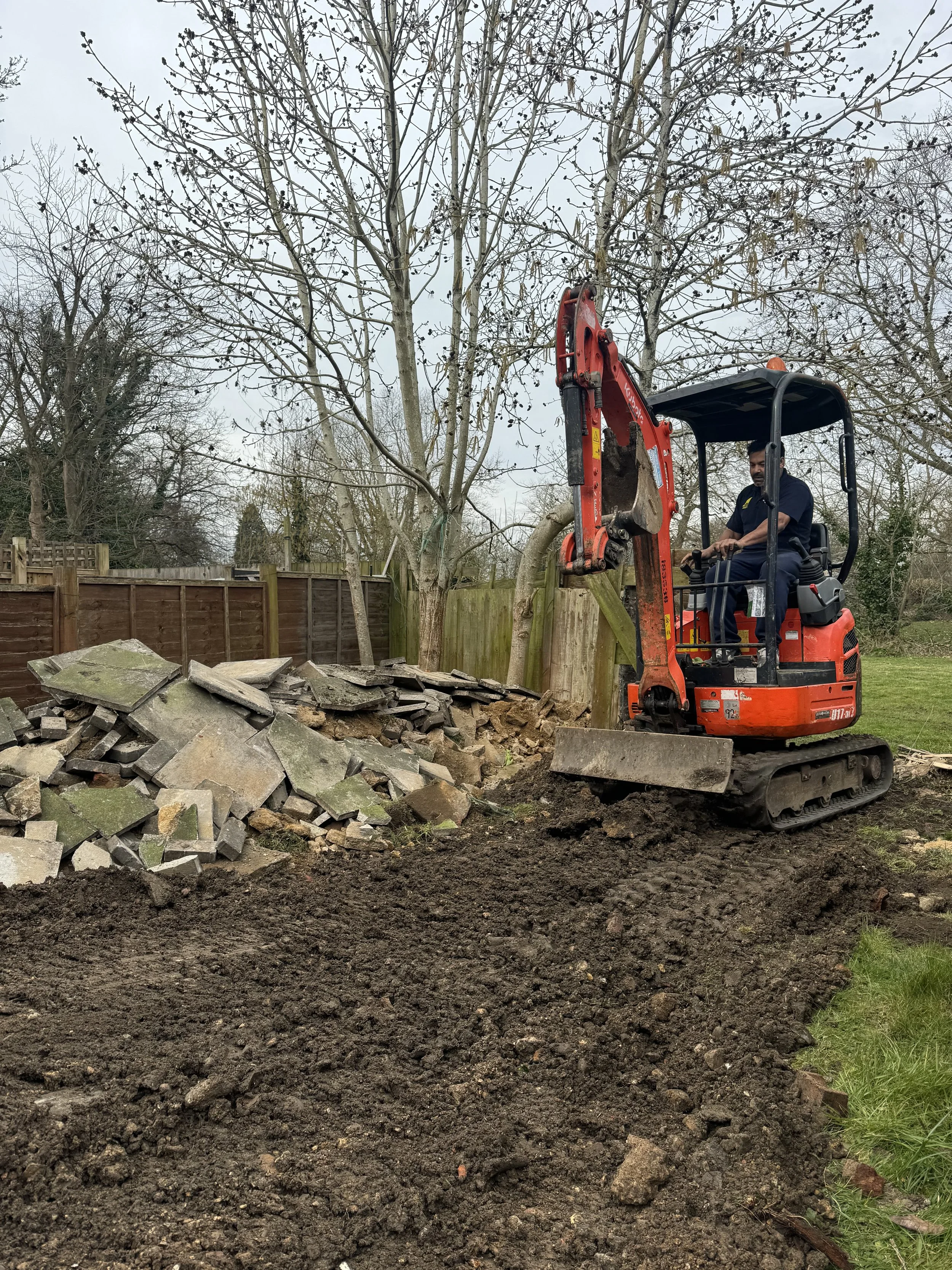 Man operating a small red excavator digging in a backyard near a wooden fence and leafless trees.
