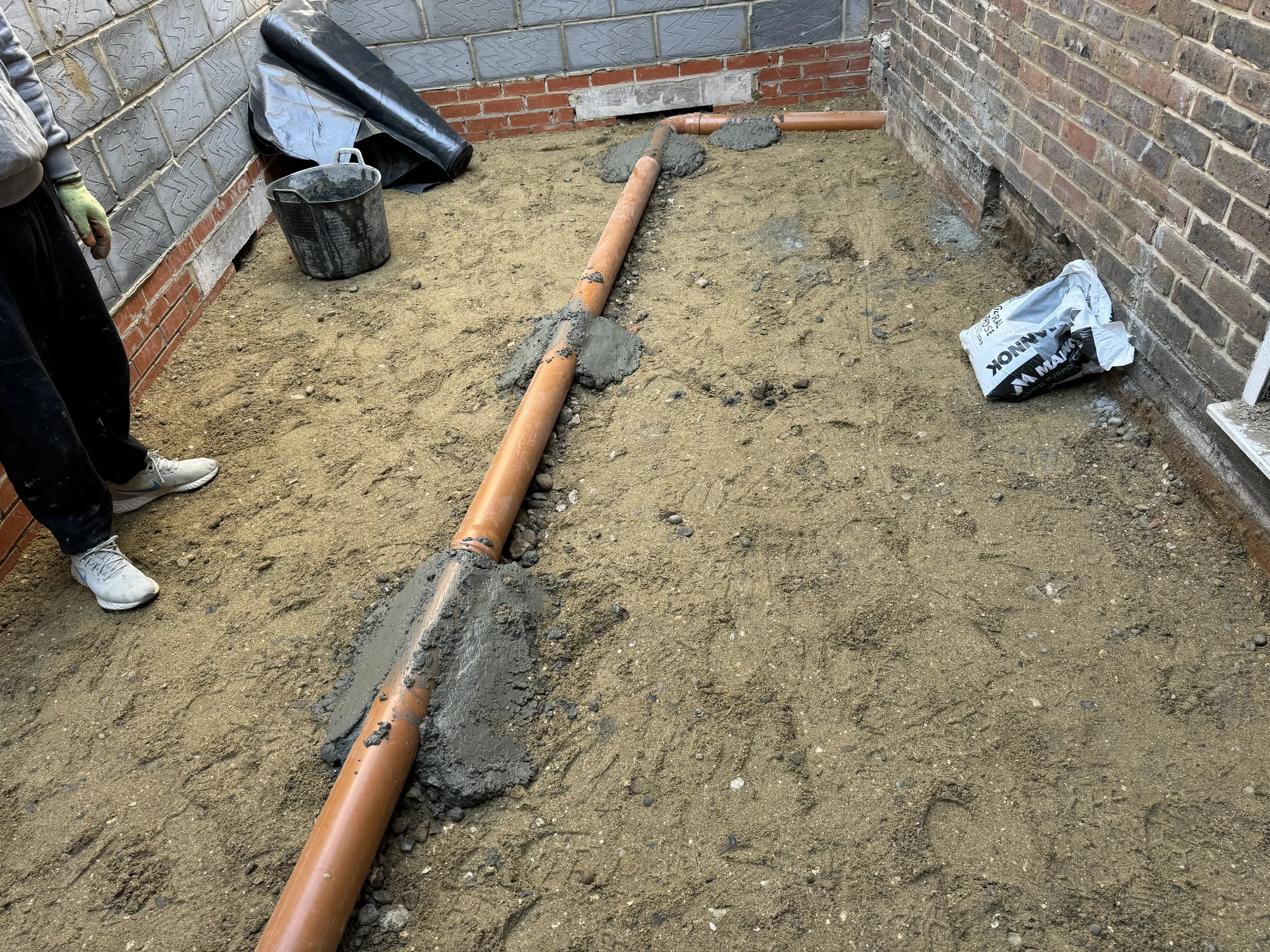 Construction site with a PVC pipe laying on a bed of sand, surrounded by construction materials and tools, with two brick walls on each side.