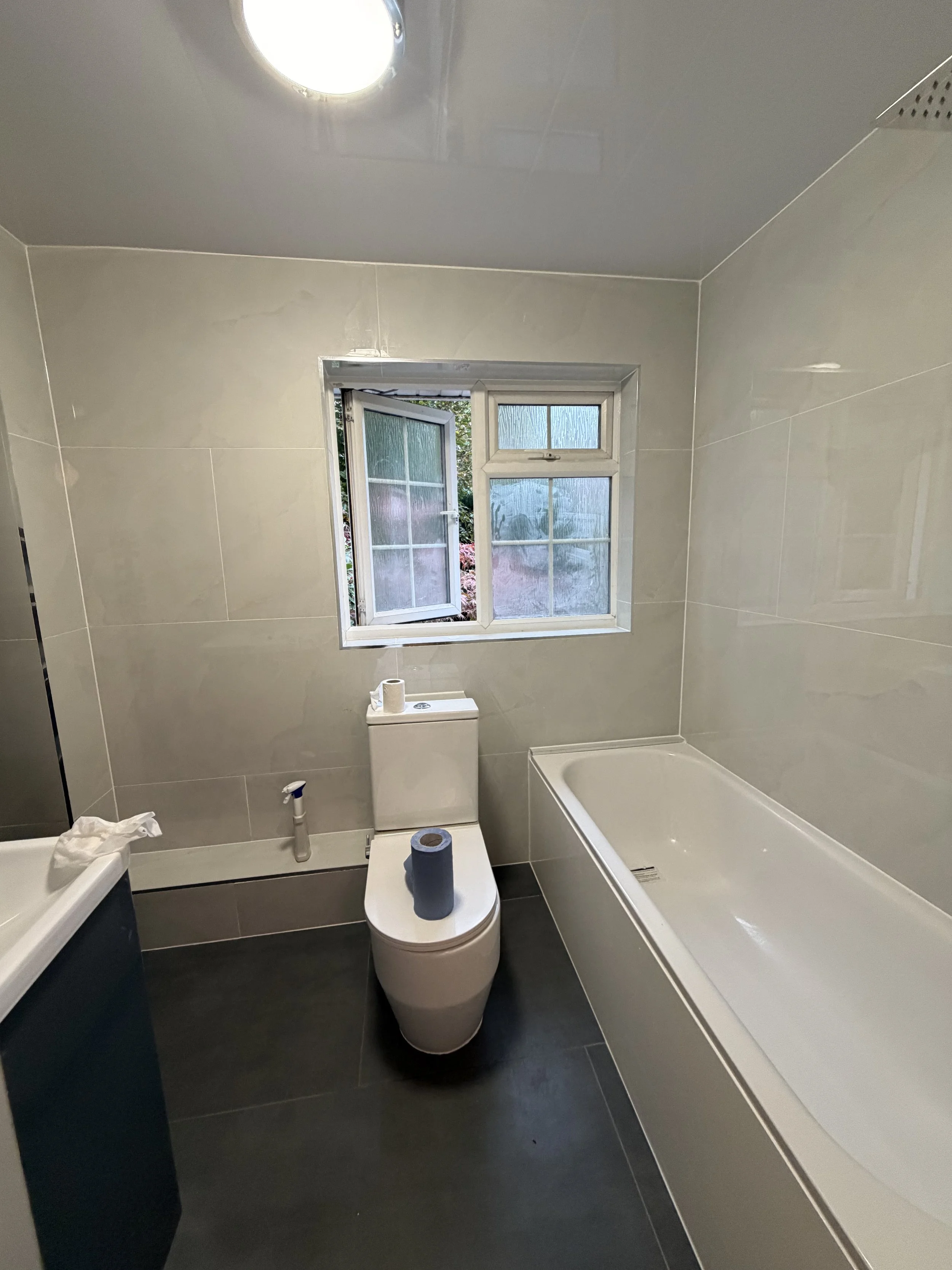 A small bathroom with a toilet, bathtub, and a window with four panes, one of which is open. There is a roll of toilet paper on the toilet tank and a roll of blue paper on the toilet seat. The bathroom has beige tiled walls and a black floor.