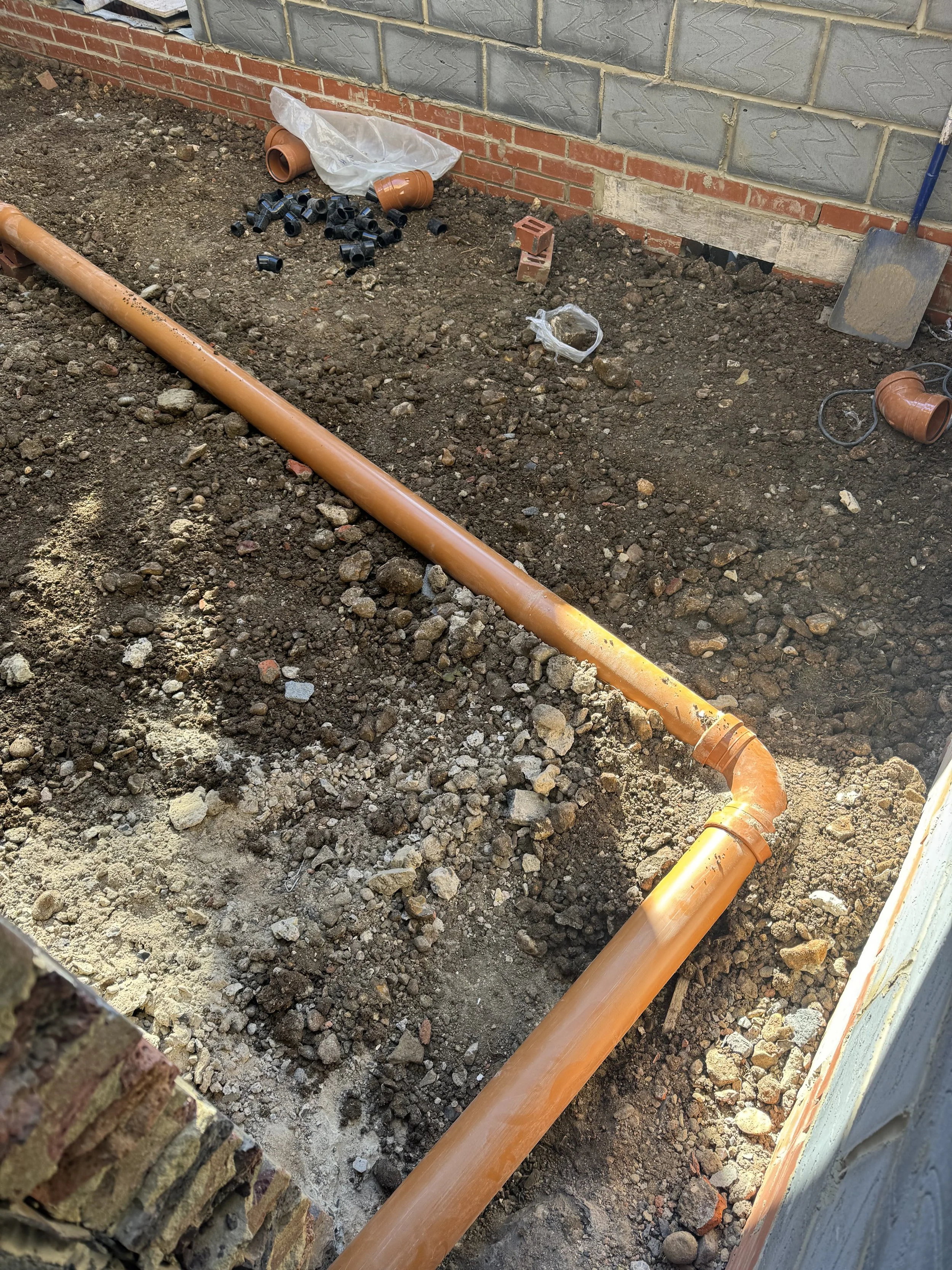 Construction site with orange PVC pipes, dirt, and loose rocks in front of a brick wall, with construction tools and materials scattered around.