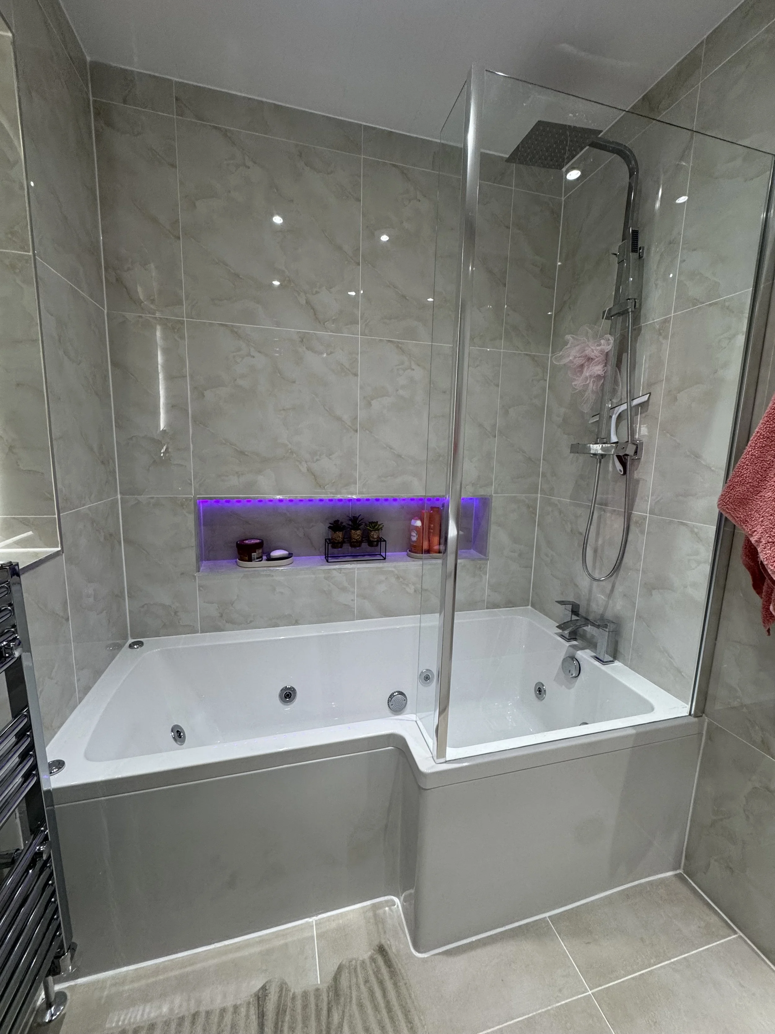 Modern bathroom with a combined bathtub and shower, marble tiled walls, built-in shelf with purple LED lighting, and a glass shower partition.