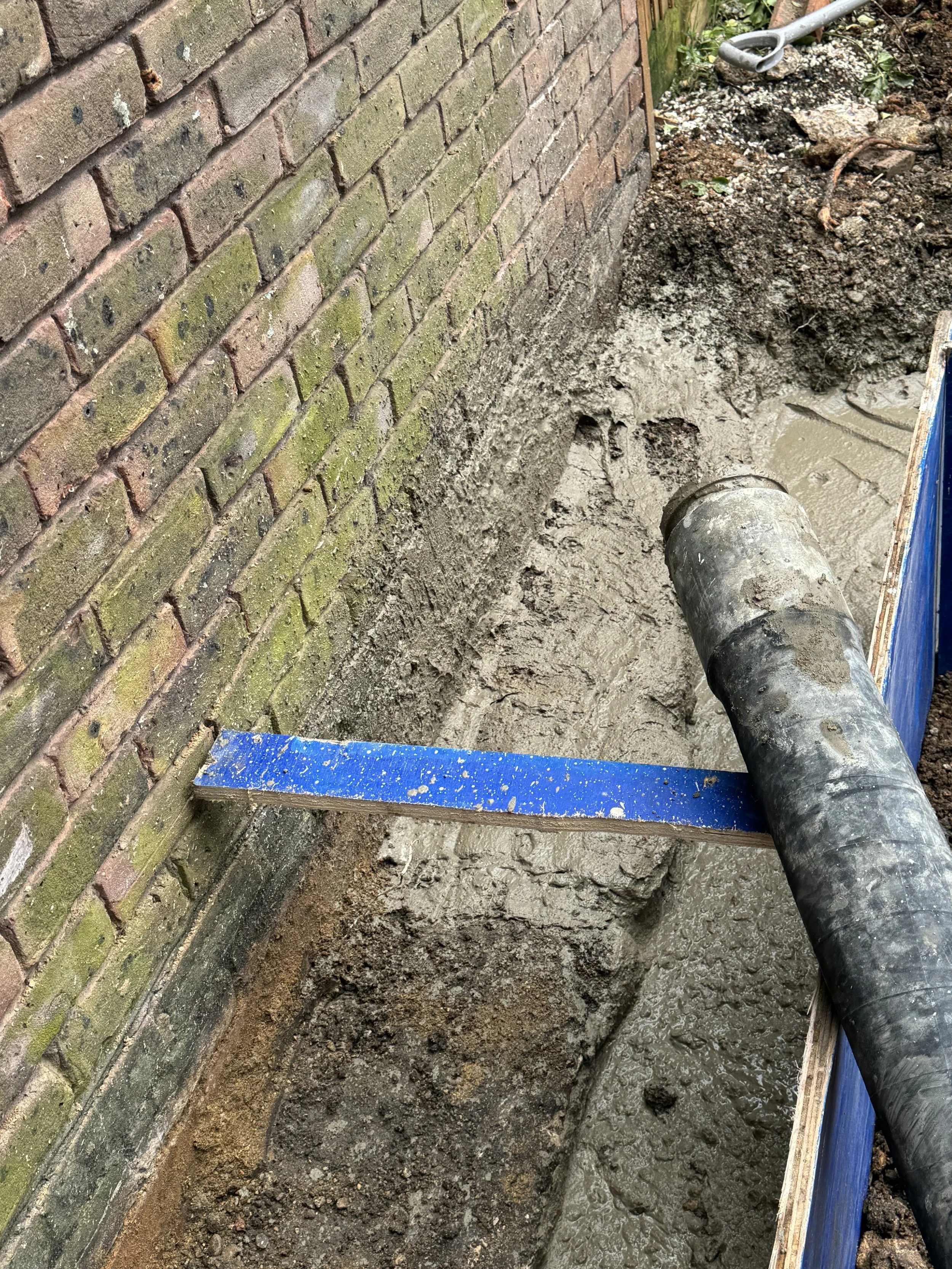 Construction site showing a brick wall and a trench being filled with wet concrete with a large black pipe and wooden formwork.