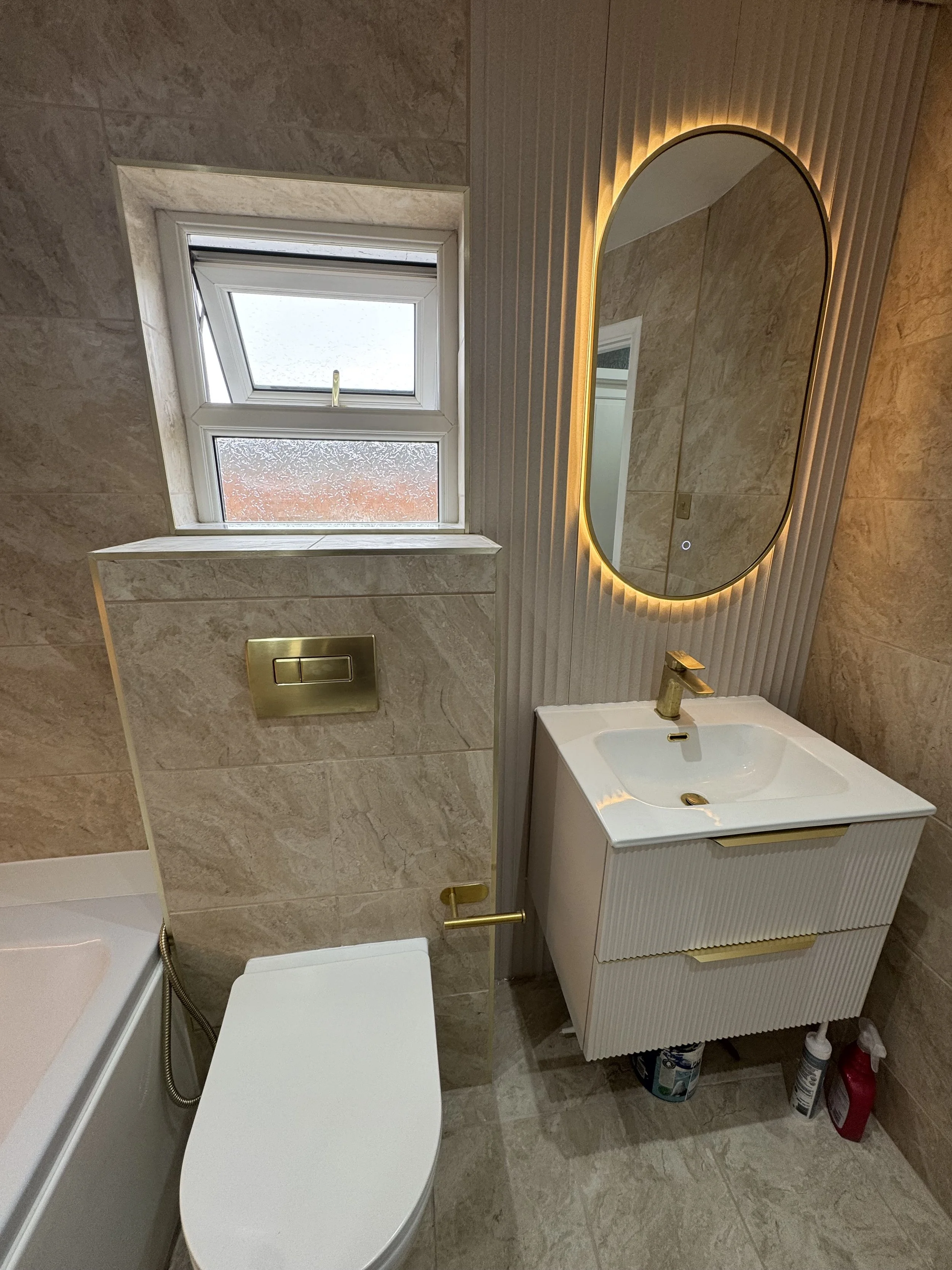Modern bathroom with beige tiled walls and floors, a small window with frosted glass, an oval backlit mirror, a white vanity with a gold faucet, and cleaning supplies underneath.