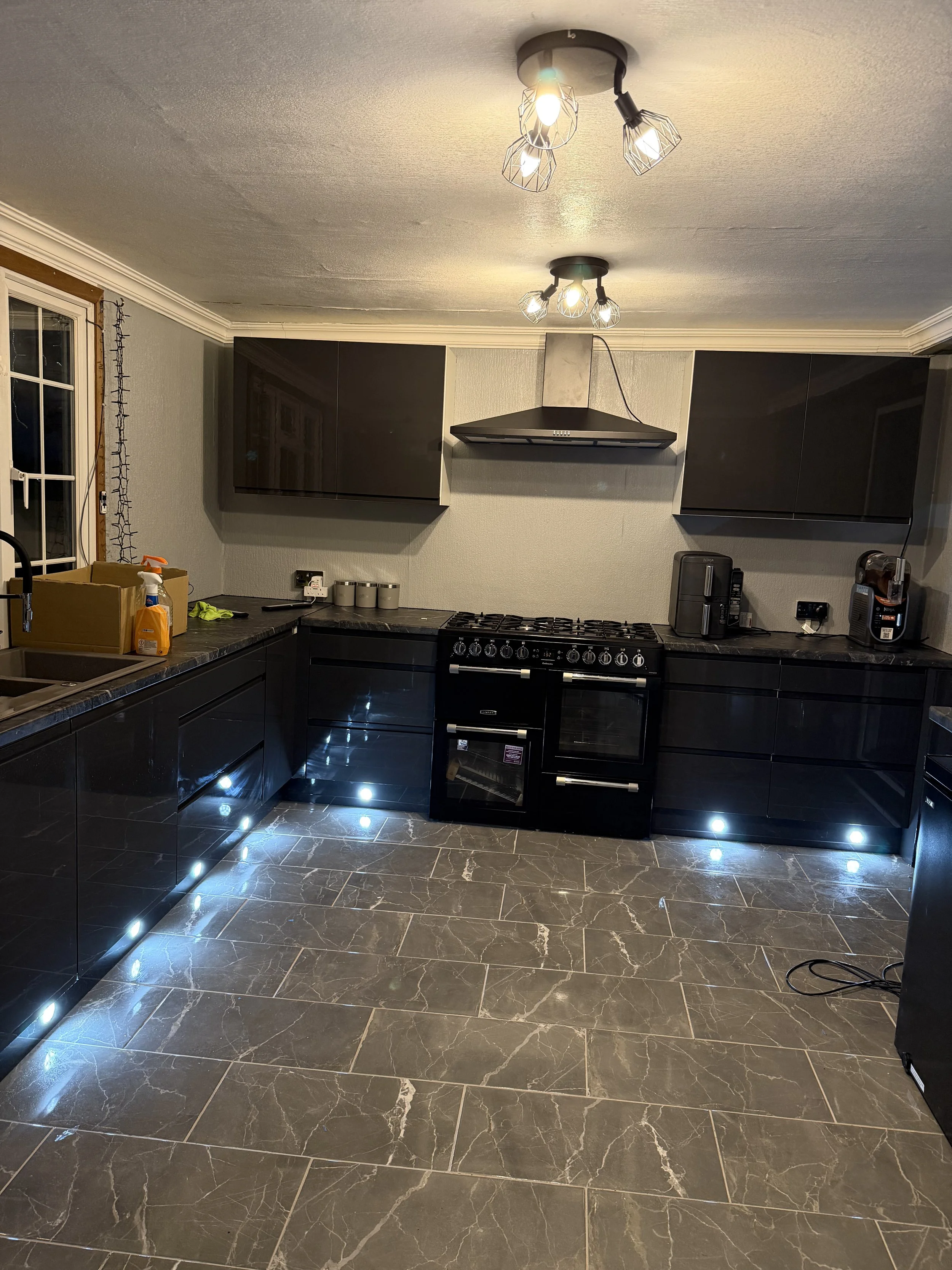 Modern kitchen with black cabinets, black stove, and black appliances, illuminated by ceiling and under-cabinet lighting, with gray tiled flooring and a window with string lights.