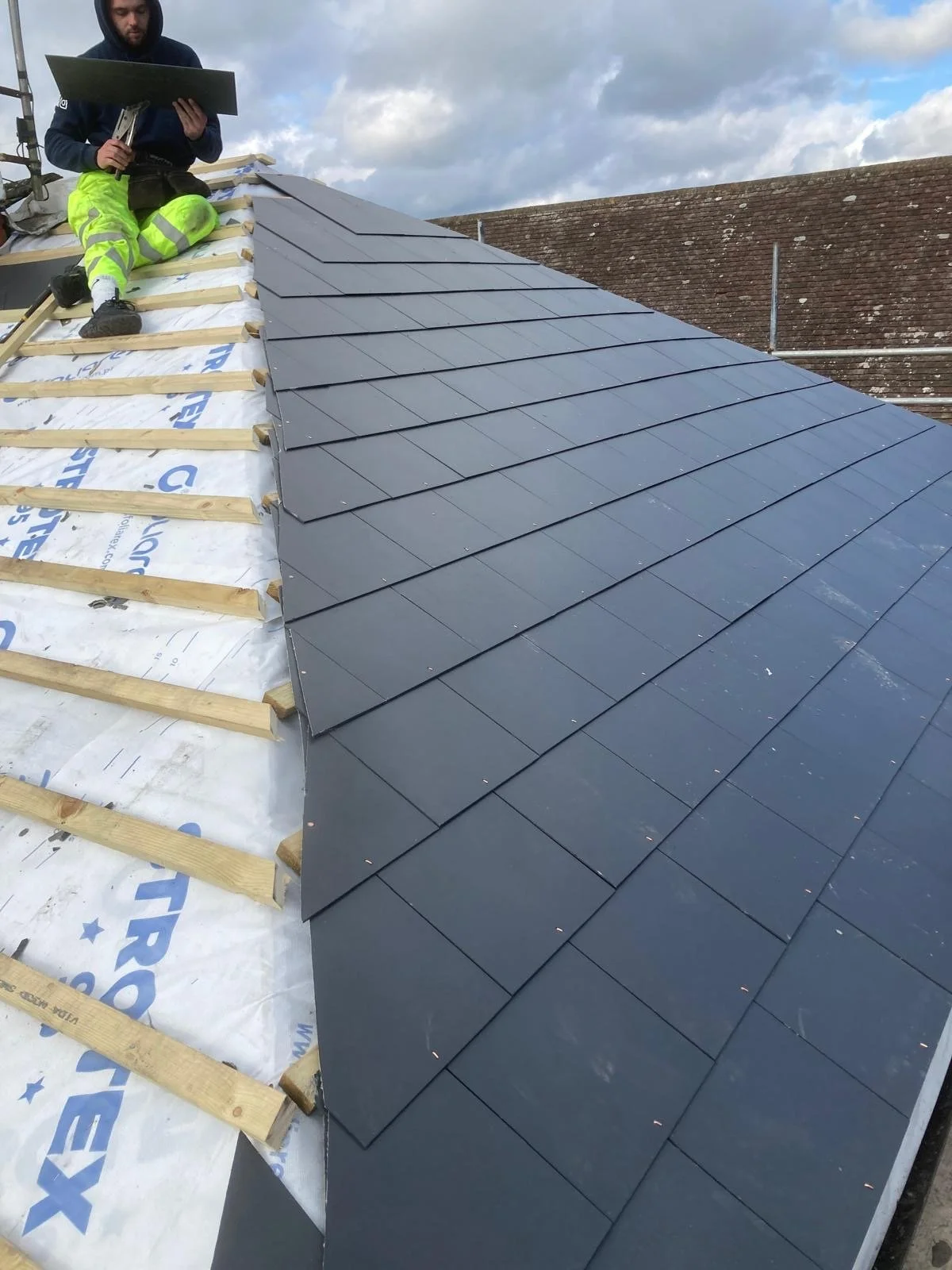 A construction worker sitting on a roof, installing dark gray shingles. He is wearing high-visibility yellow pants and a dark hoodie. The roof is partially covered with shingles, and the sky is cloudy.