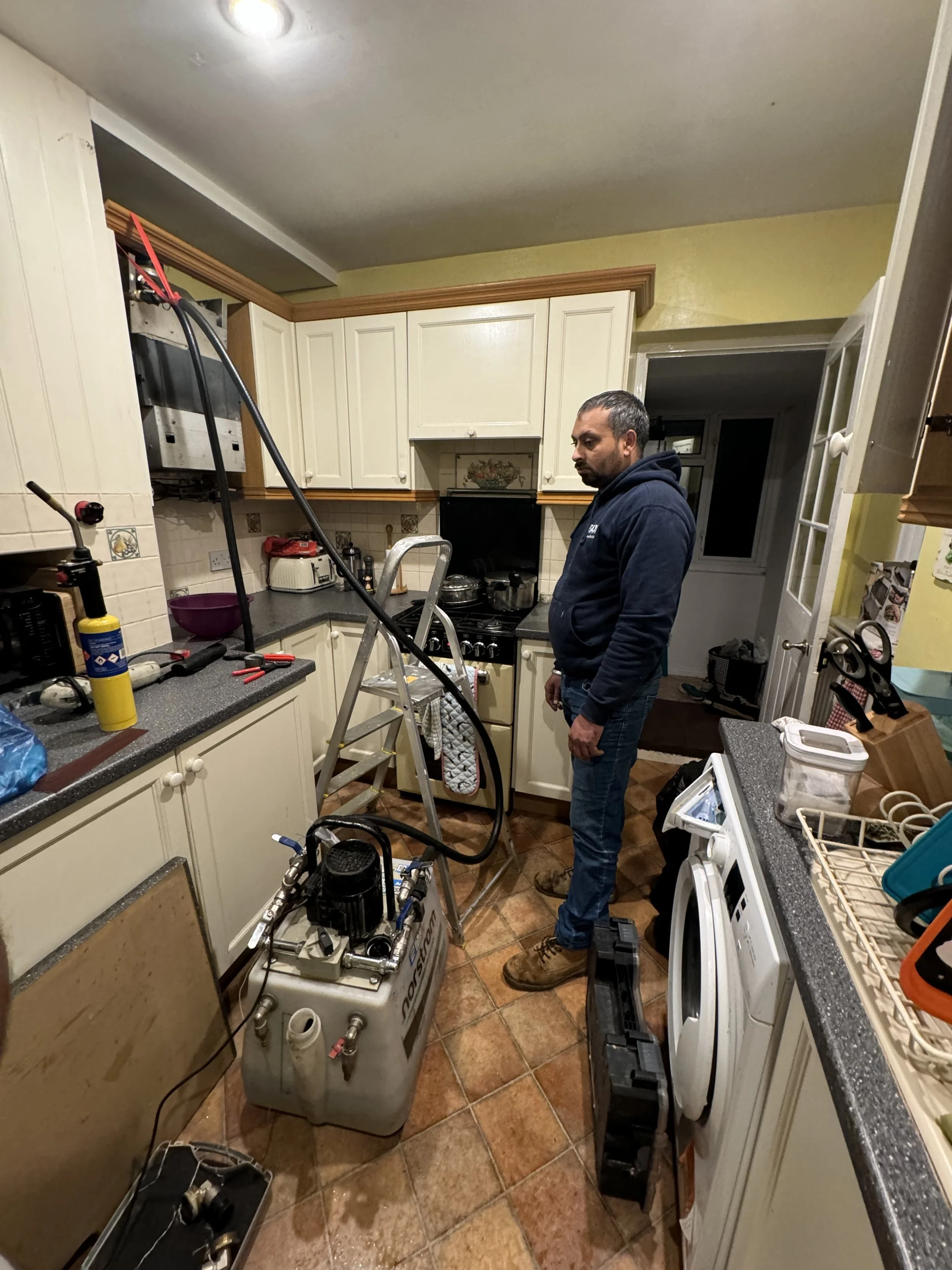 Man in a kitchen with plumbing or HVAC equipment, ladder, and tools, working or inspecting the setup.