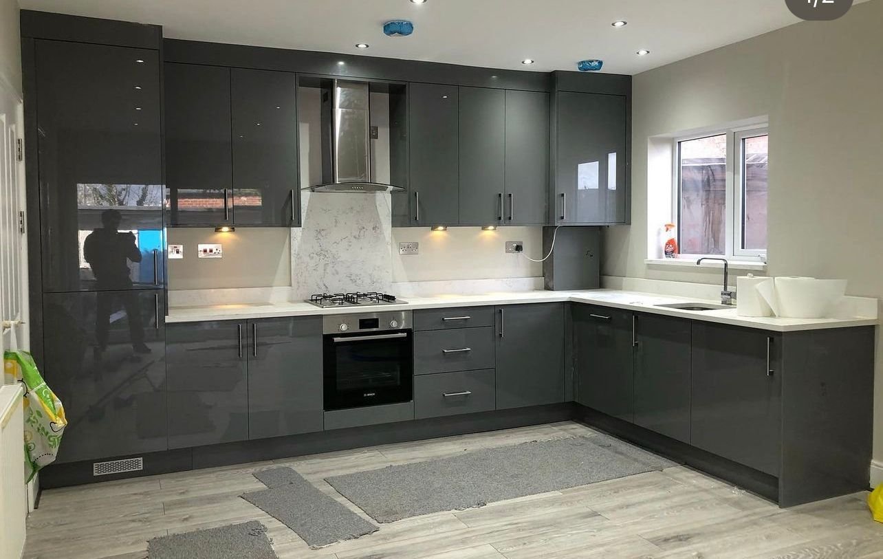 Modern kitchen with grey cabinets, white countertops, a window above the sink, and a built-in oven and gas stove. The floor is wooden with grey rugs, and there are recessed ceiling lights.