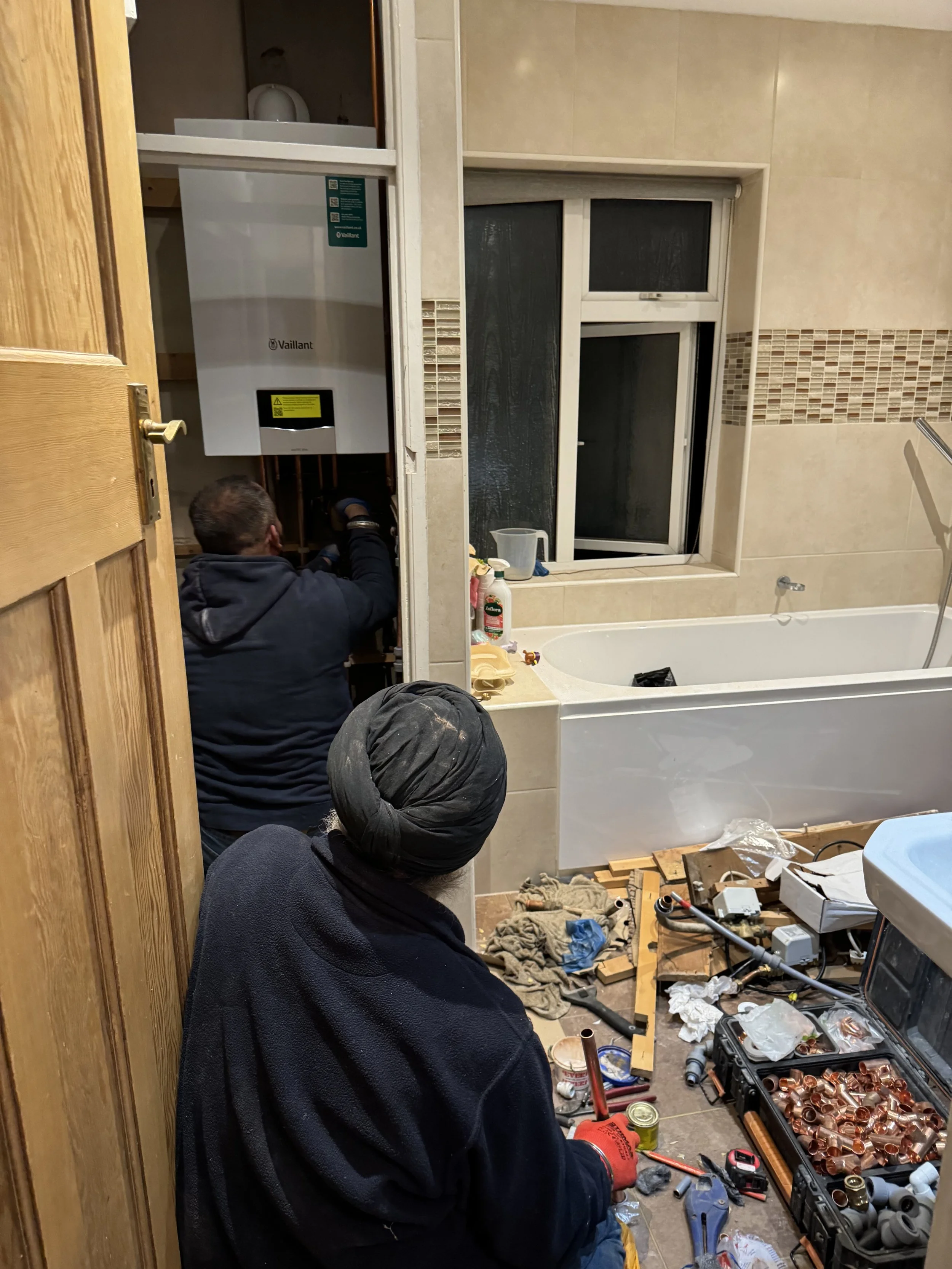 Two workers installing or repairing a boiler in a bathroom, with tools and parts on the floor.
