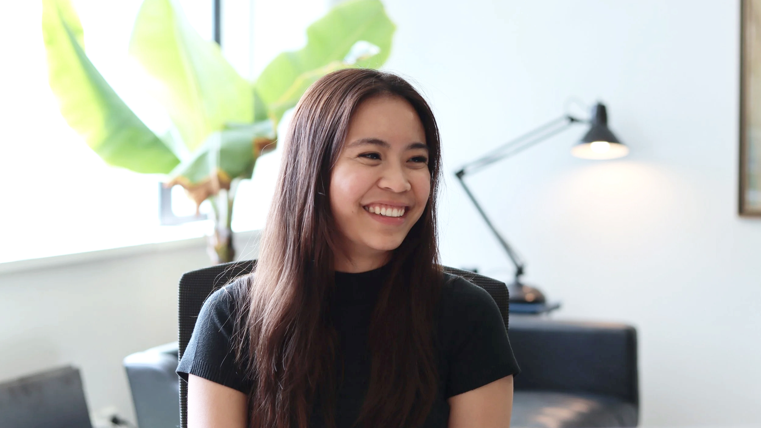 A woman with long dark hair smiling in an office setting, with a large plant and a desk lamp in the background.