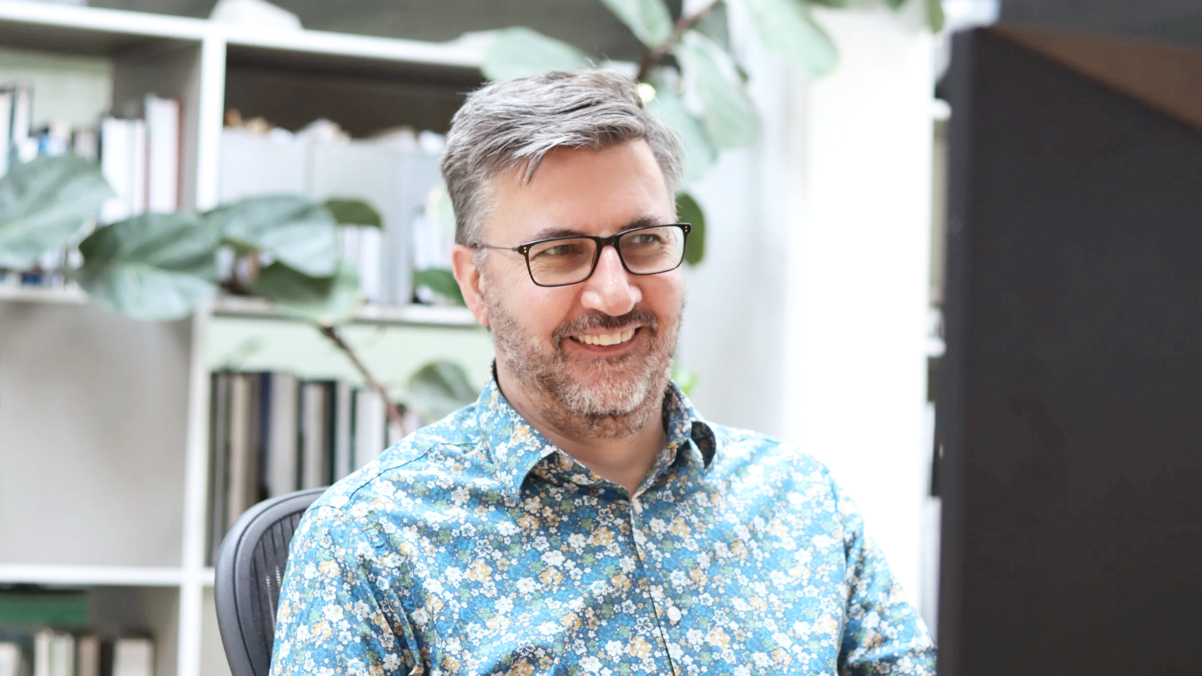 A man with glasses and a beard, smiling while looking at a computer screen. He is wearing a blue floral shirt and is seated in an office environment with books and large green plants in the background.