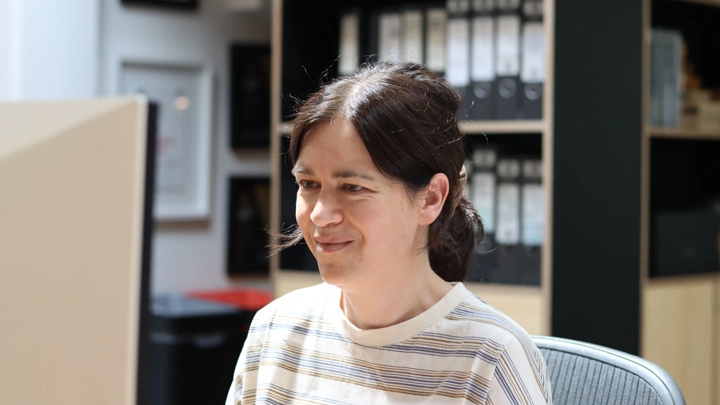 A woman with dark hair sitting at a desk, looking at a computer screen with a slight smile, in an office environment with shelves and binders in the background.