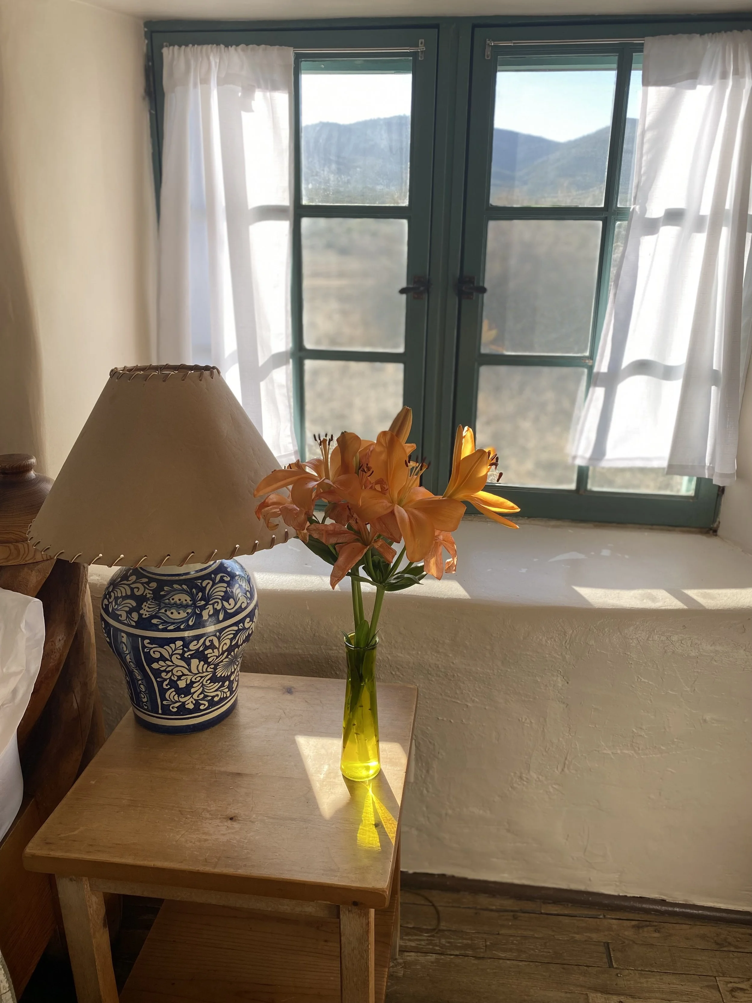 A side table carrying a beautiful ornamental lamp stump with a sutured leather lamp cover and bright orange lilies dress the area with color. Behind are teal juliet window panes, draped in white linen curtains.