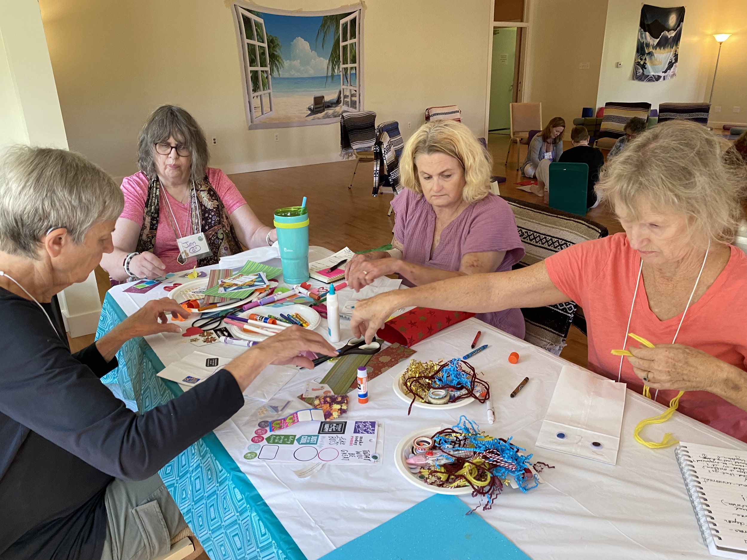 Group of people gathered around a table working on arts and crafts, with colorful supplies such as markers, glue, and ribbons, in a room decorated with beach-themed art and cozy seating.