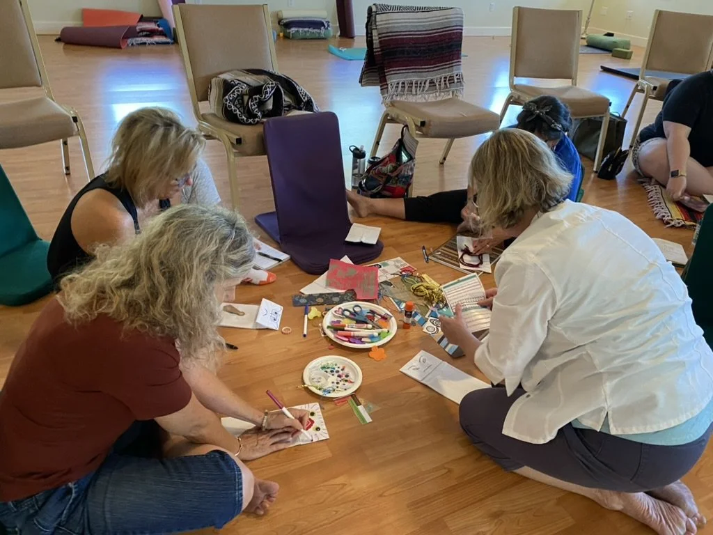 Group of women sitting on the floor engaging in arts and crafts, with various supplies like markers, paper, and scissors, in a room with chairs and a wooden floor.