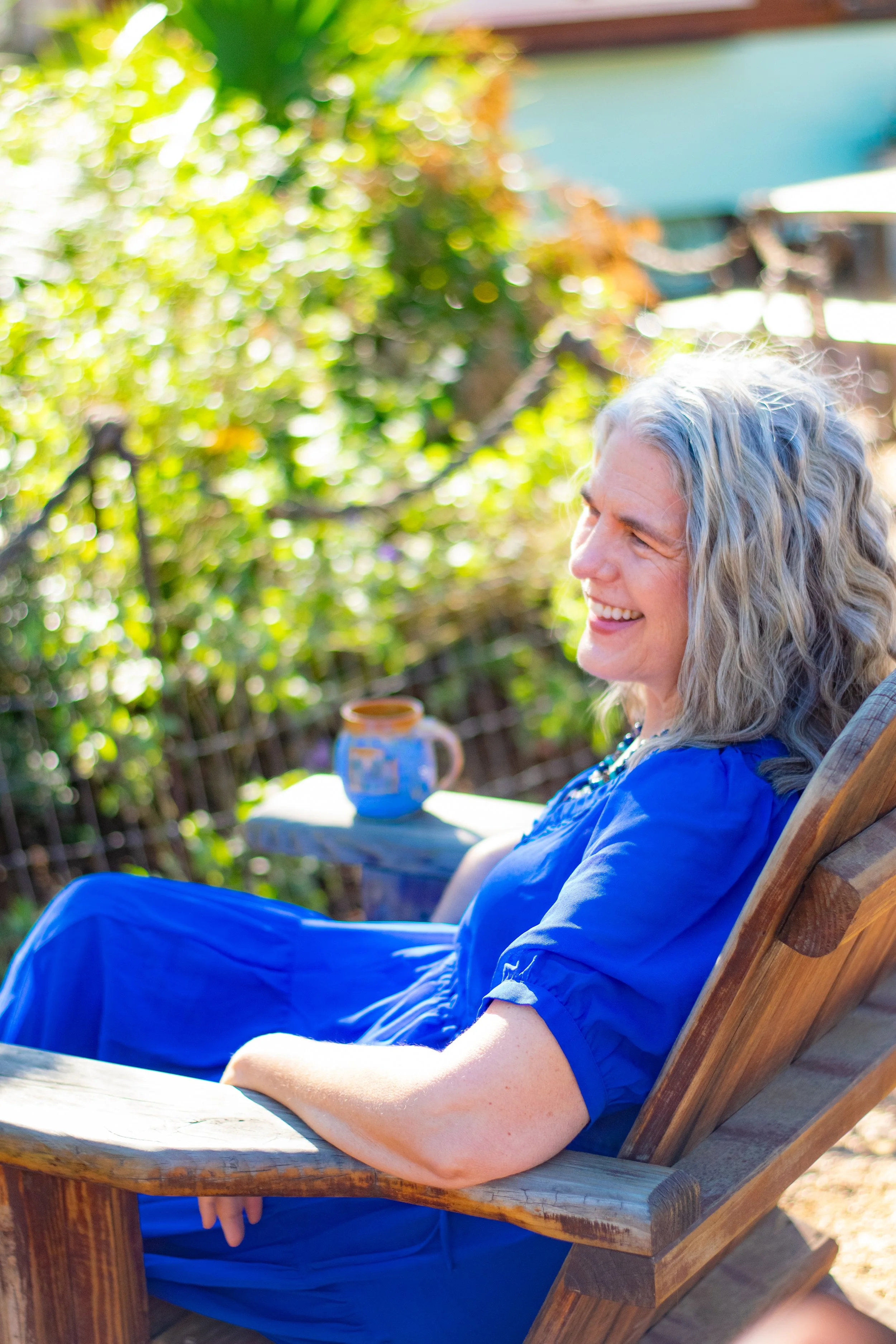 Carolyn, the founder and guide of these services sits on an adirondack chair in a bright blue maxi dress accompanied by her blue cermaic coffee mug on the arm rest..