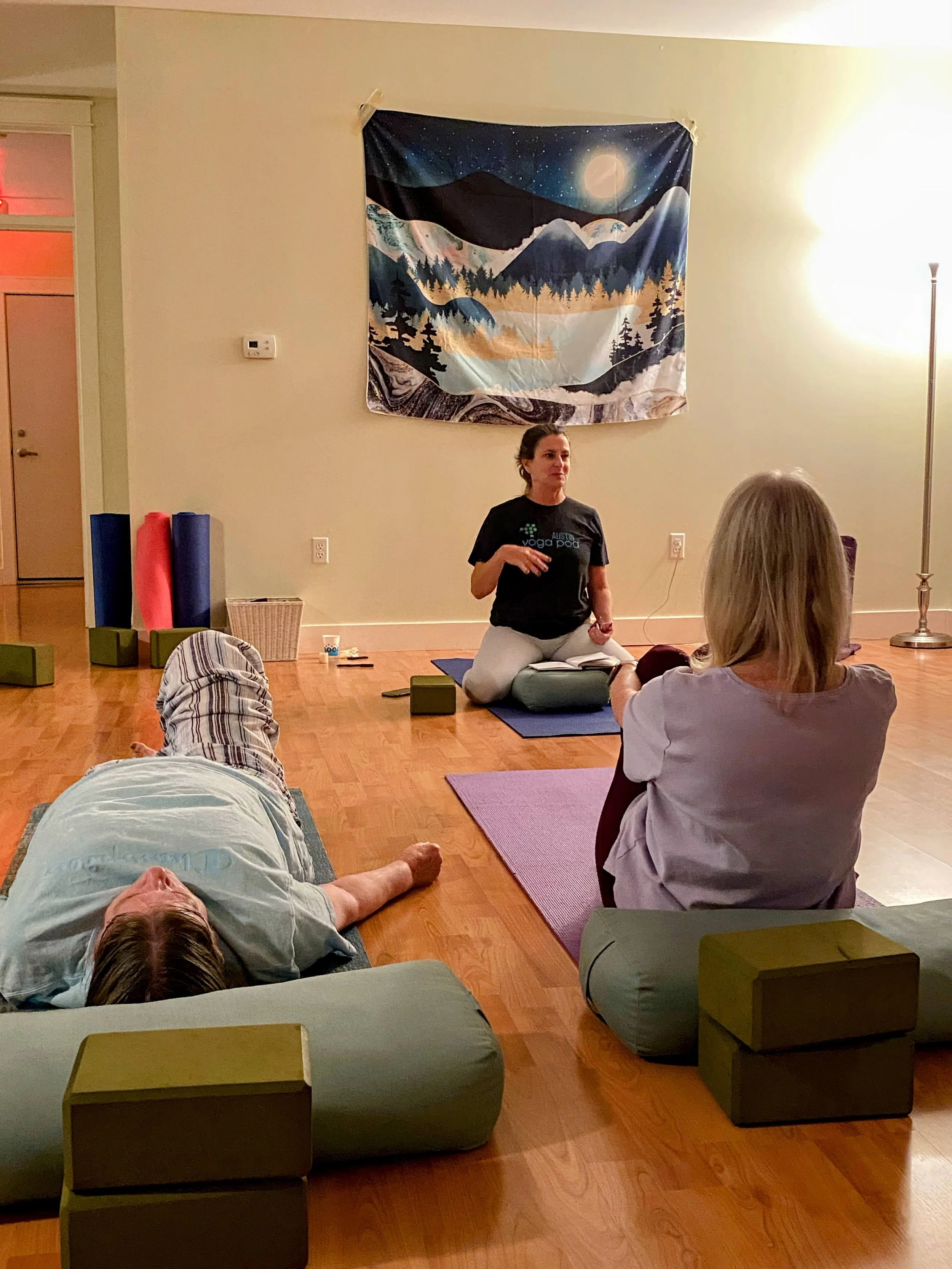Women on their yoga mats during a guided yoga meditation. The instructor is speaking instructionally to the class as they relax and listen.