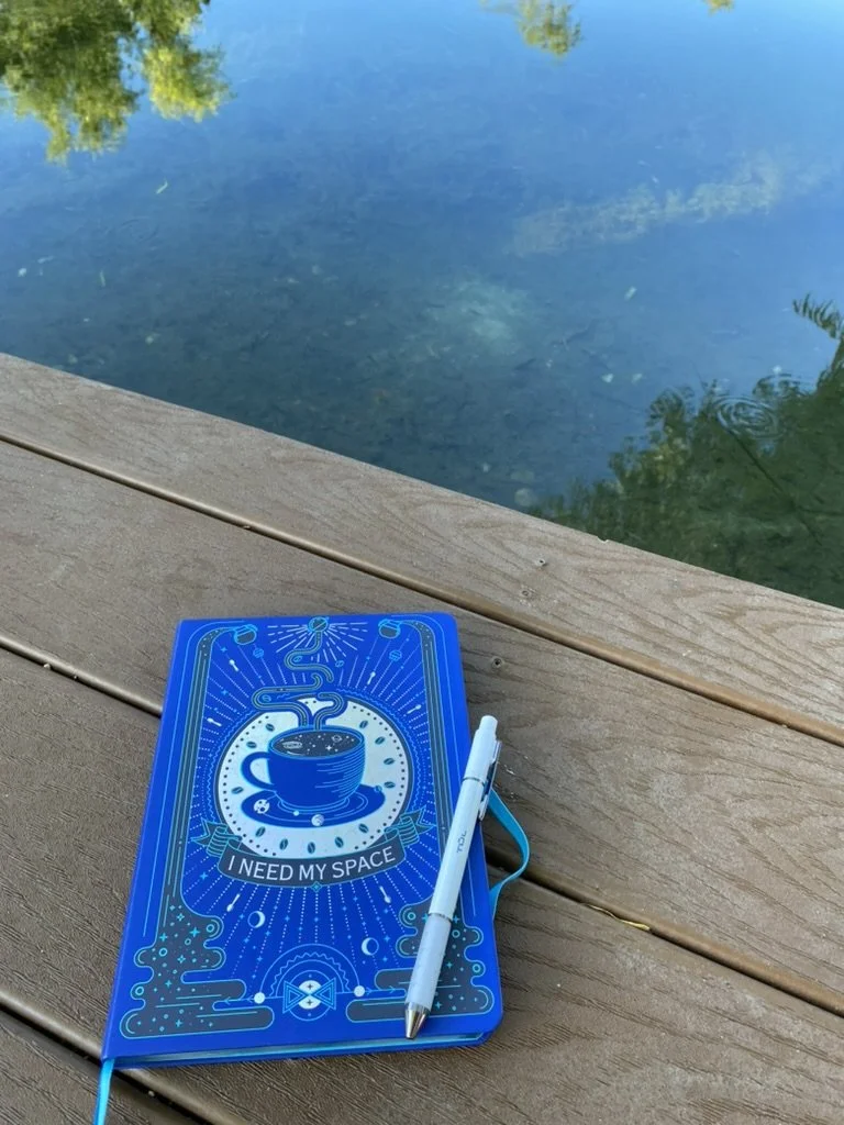 A blue notebook with a space-themed design and the phrase "I NEED MY SPACE" on a wooden deck near a body of water, with a white pen attached to it.
