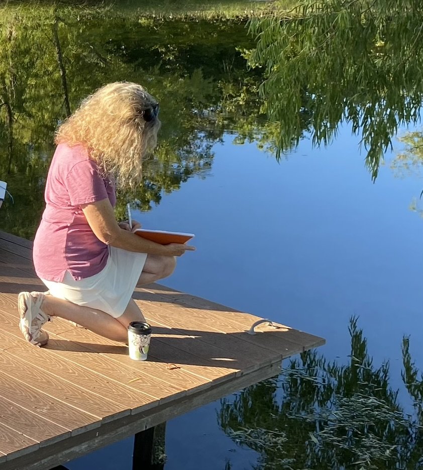 A woman with curly blonde hair, wearing a pink shirt and white skirt, kneeling on a wooden dock while reading a book by a calm body of water surrounded by green trees.