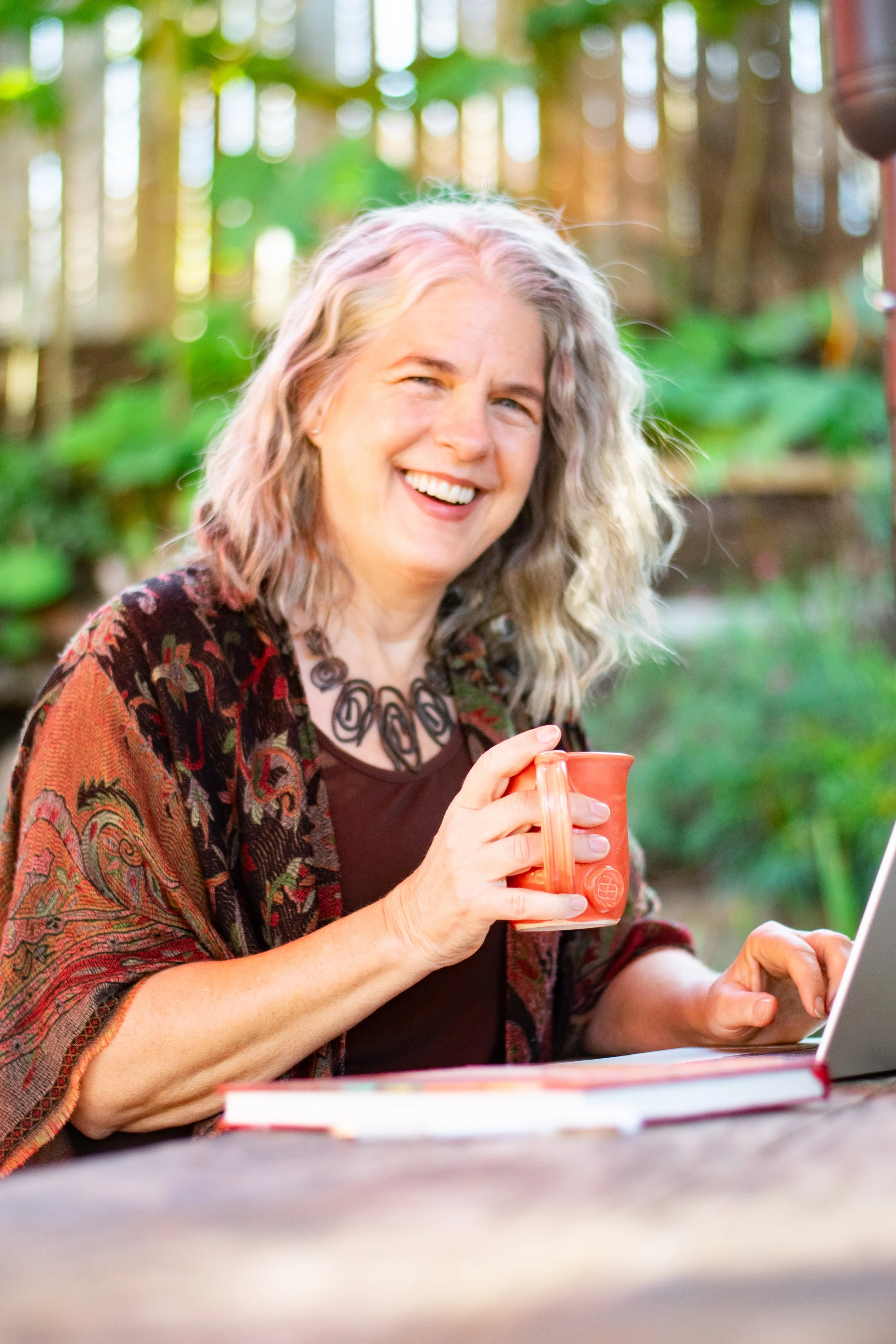 Candid photo of Carolyn glancing at camera while on her laptop and sipping on a coffee, surrounded by green, lush, nature.