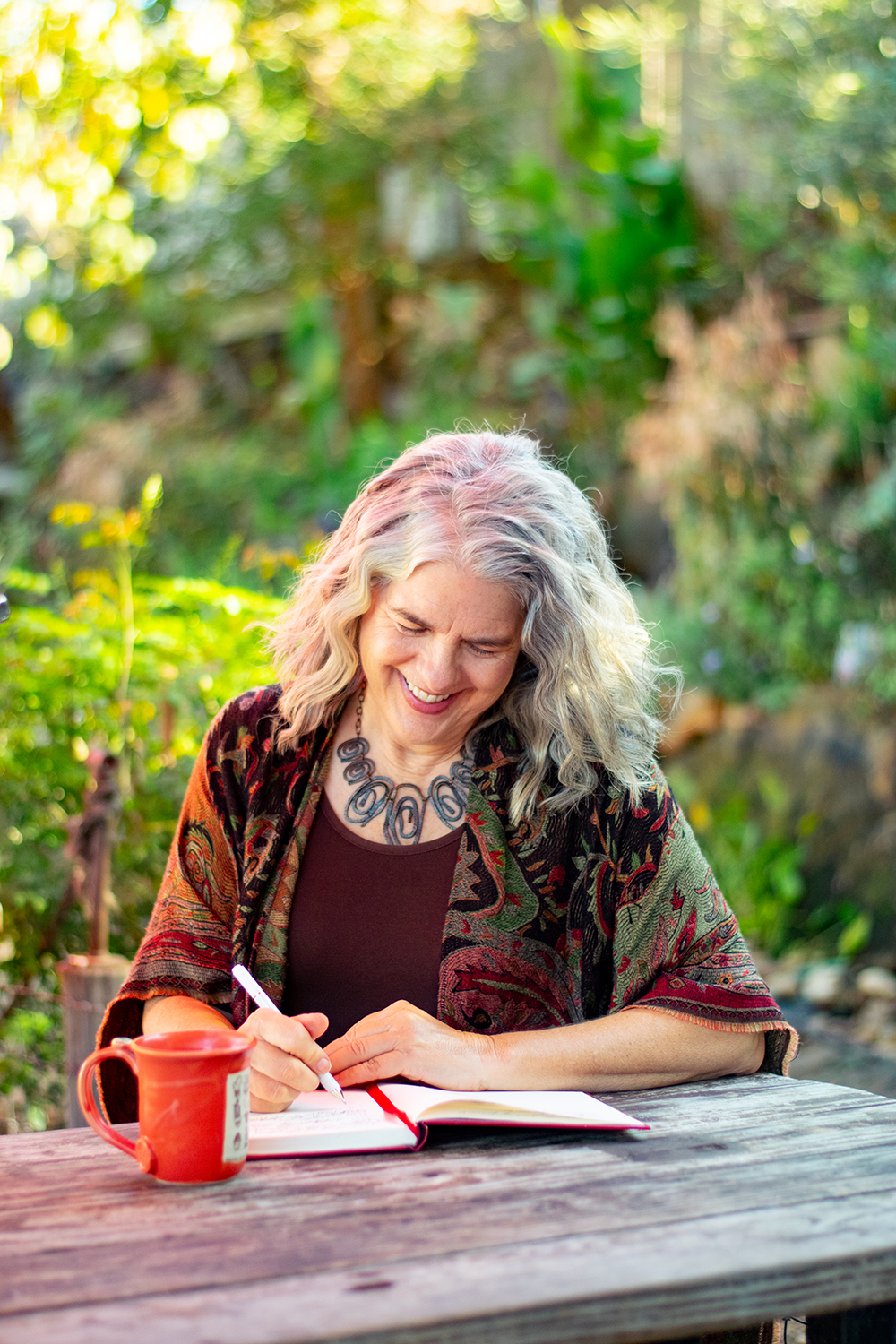 Carolyn, sitting outside on a picnic bench at a coffee shop, looks down at her journal contently as she writes.
