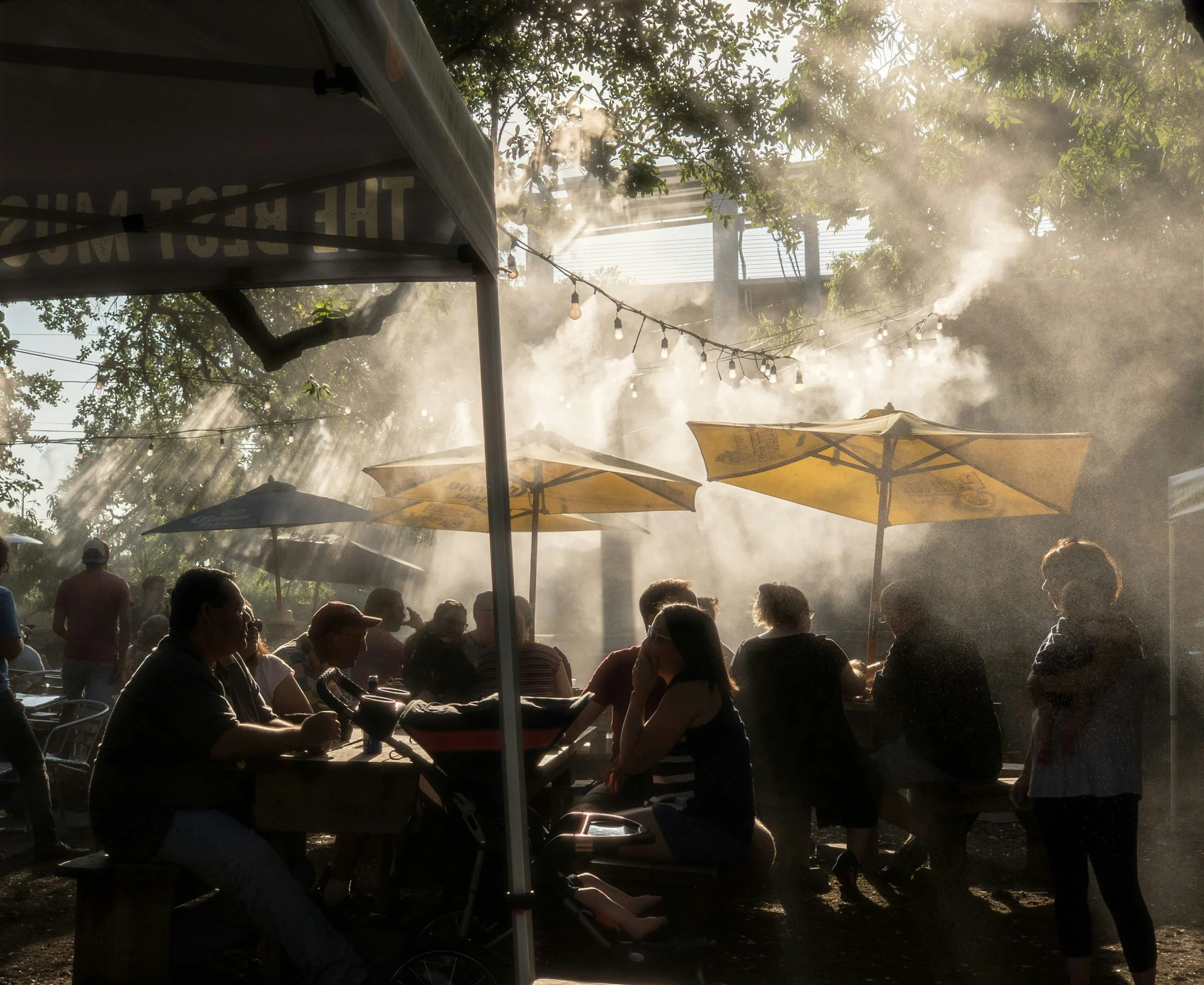 Mist and yellow umbrellas cover groups of people sitting outside to cool everyone off in the low Texas heat. Location is unknown.