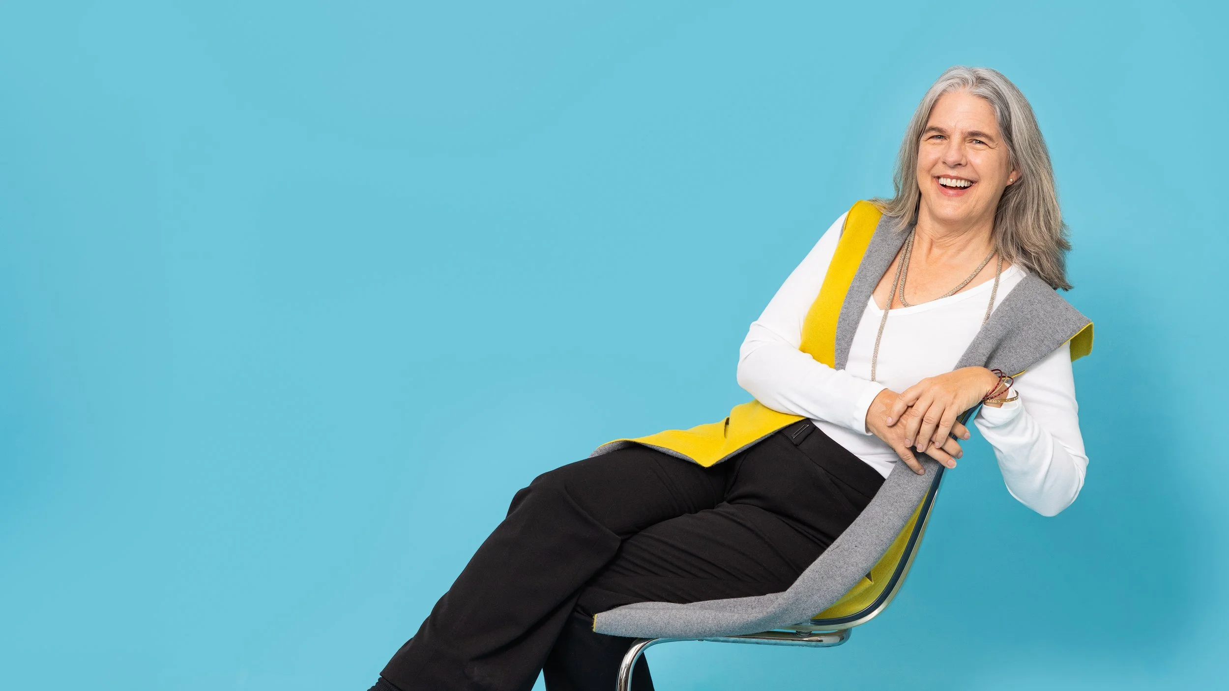 Carolyn playfully leaning on a modern looking chair, with a yellow vest on, surrounded by a baby blue backdrop.