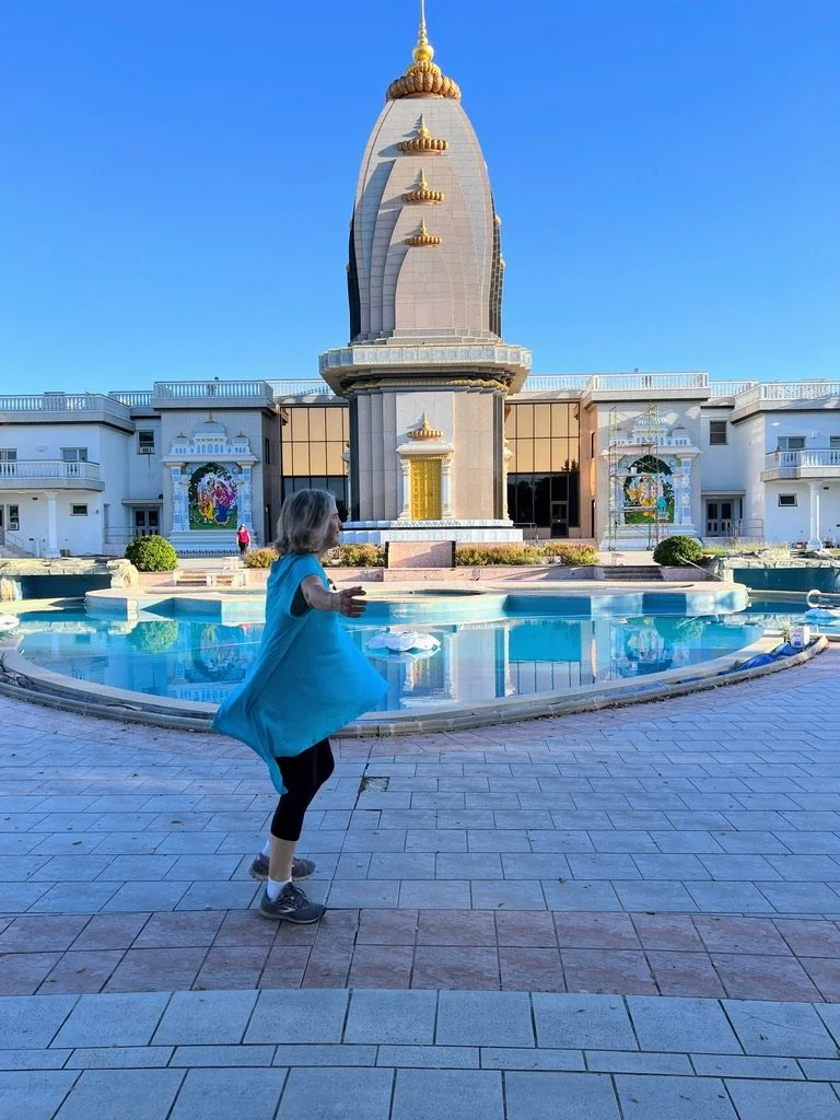 A young girl wearing a blue dress and black leggings playing near a fountain in front of a Hindu temple with a tall, ornate tower. The temple has white walls and colorful artwork on the sides, with a clear blue sky overhead.