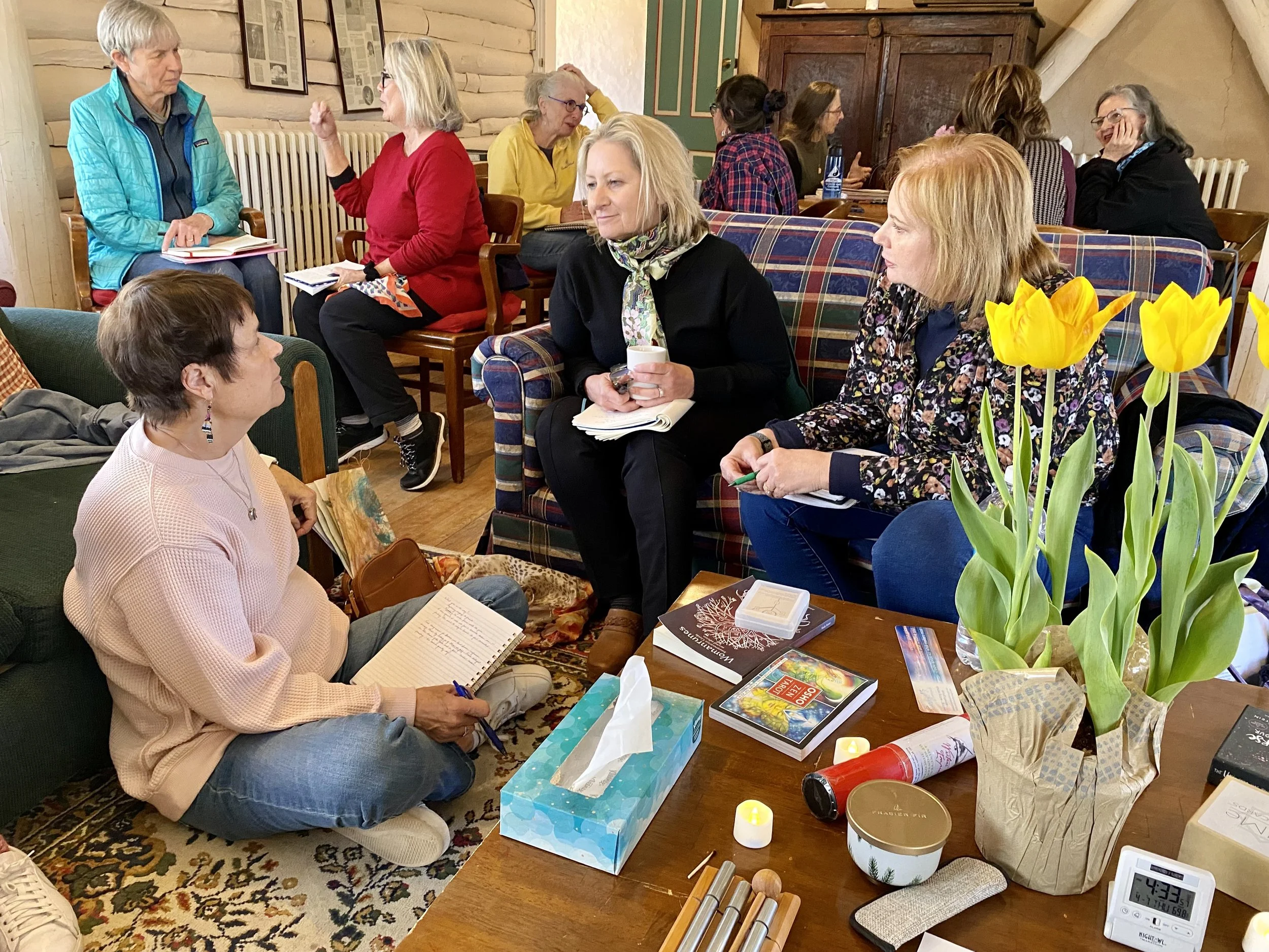 Group of three women intensely listening to each other share words. Fully bloomed yellow tulips sit to the side resembling growth in a way.