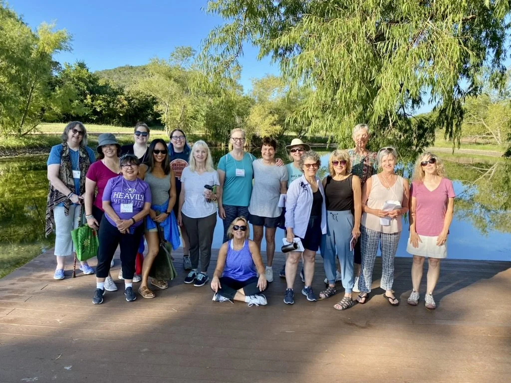Group of approximately fifteen women standing on a wooden walkway by a pond, surrounded by green trees and bushes on a bright, sunny day.