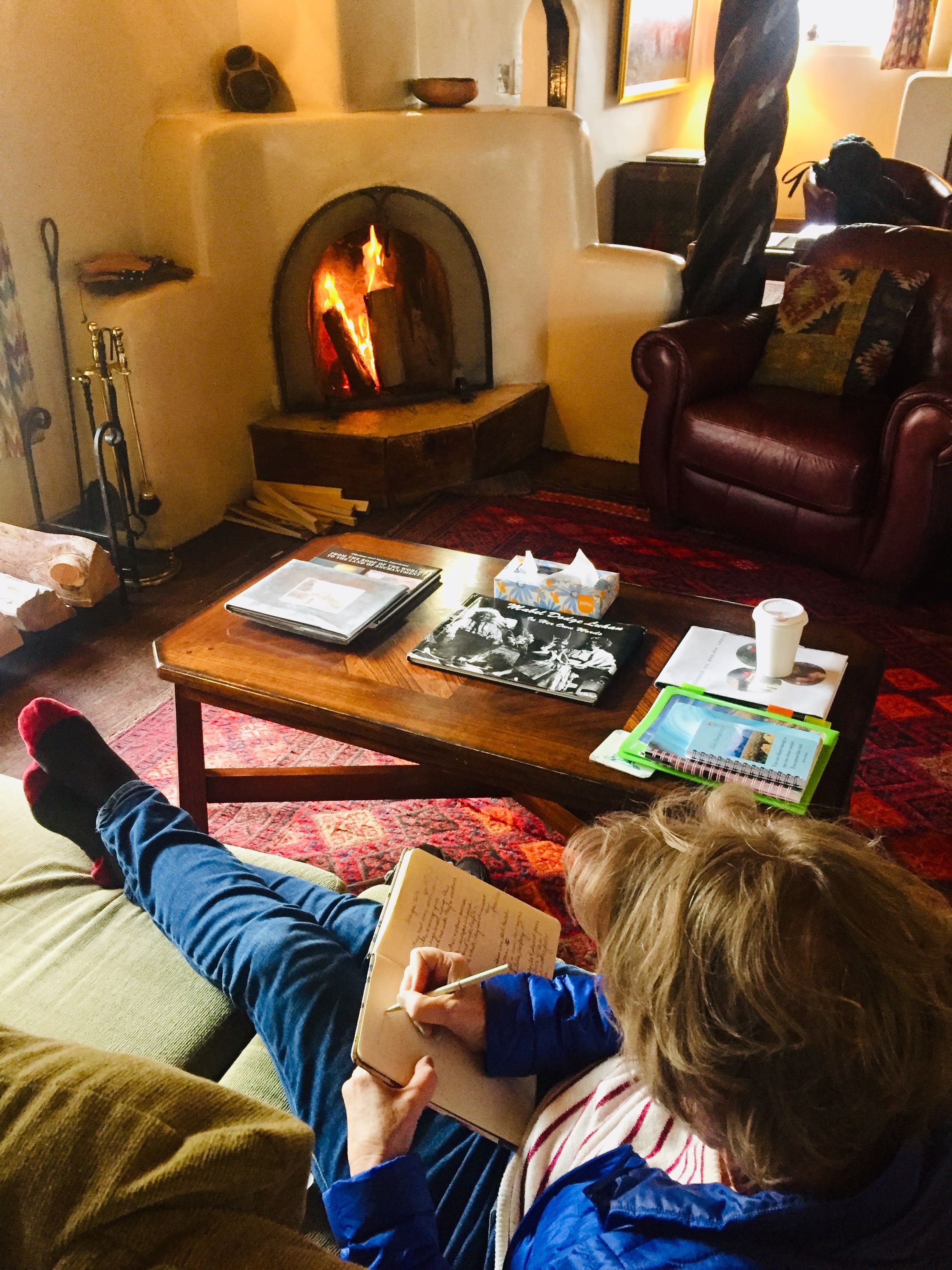 Woman with her feet up on the couch, in front of a cozy fire, writing in her journal.