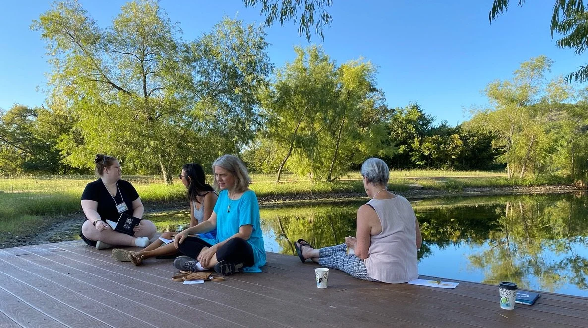 Four women sitting by a pond on a wooden deck, with trees and clear blue sky in the background, enjoying a peaceful outdoor gathering.