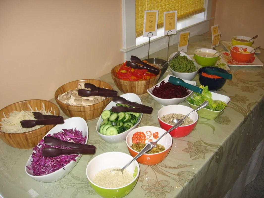 A variety of fresh vegetables and salad toppings arranged in bowls on a buffet table, including shredded cheese, shredded purple cabbage, sliced cucumbers, sliced red and yellow peppers, lettuce, shredded beets, shredded carrots, and various dressings.