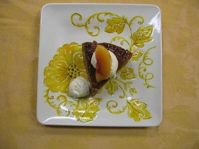 Slice of chocolate cake with a dollop of cream, topped with a peach slice, on a decorative floral plate.