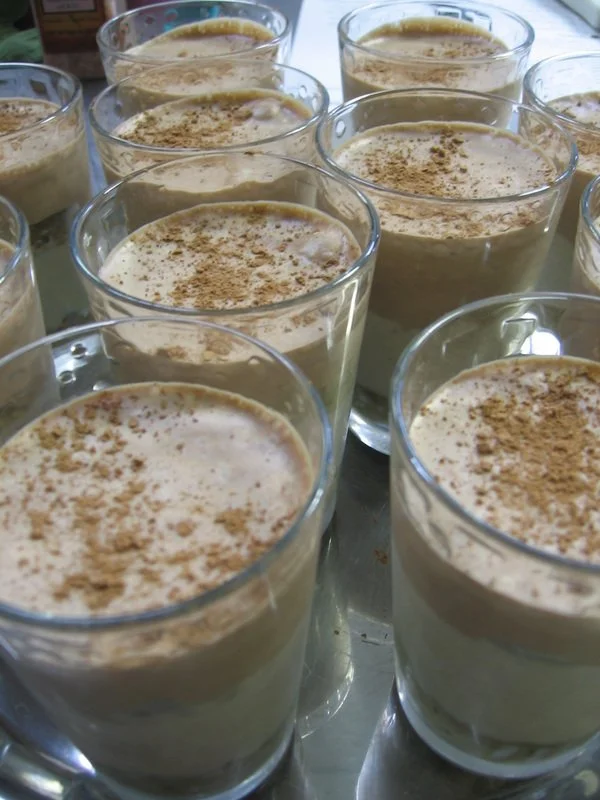 Multiple glasses of creamy dessert, possibly tiramisu or pudding, topped with a sprinkle of cinnamon or cocoa powder.