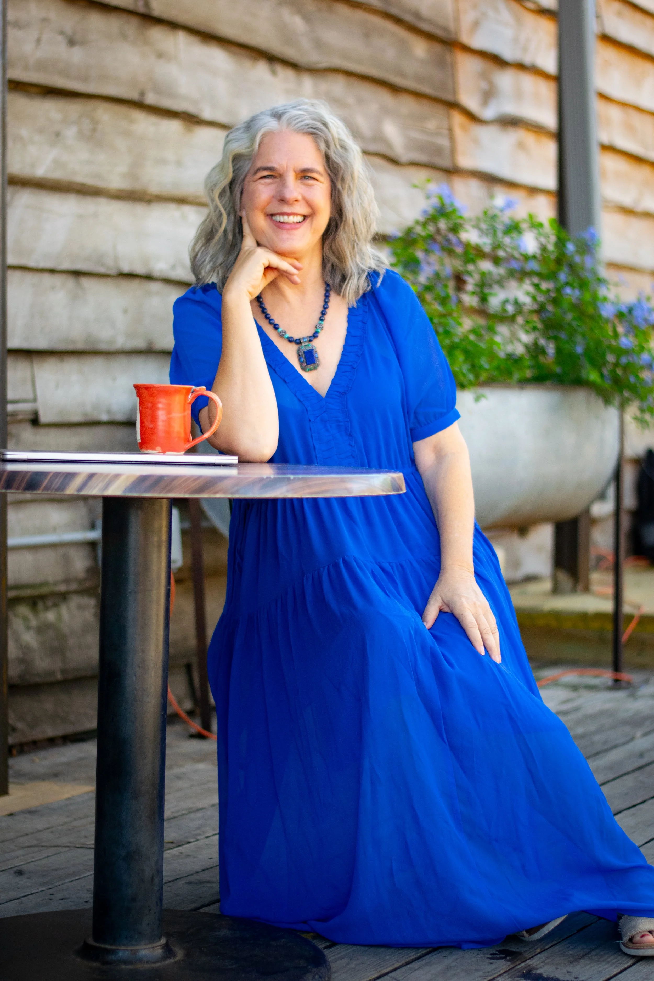 Headshot of Carolyn, with silver wavy hair, a big smile, chunky beautiful necklace, a flowy blue maxi dress, sitting by her orange ceramic coffee cup.