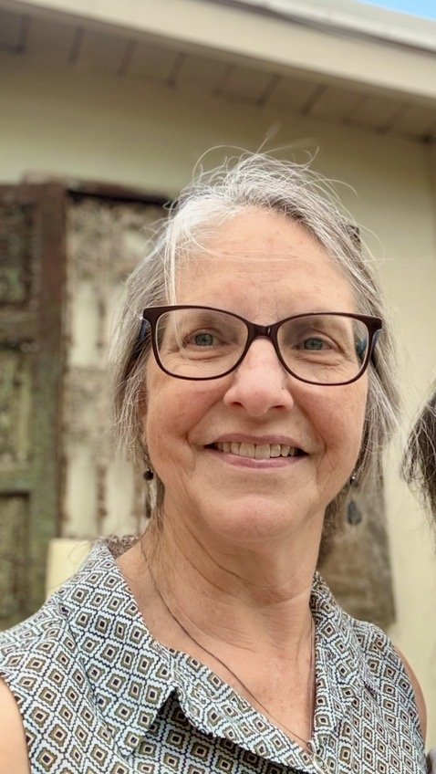 Smiling older woman with glasses and gray hair, wearing a patterned sleeveless blouse, outdoors in front of a building with a fence and trees.