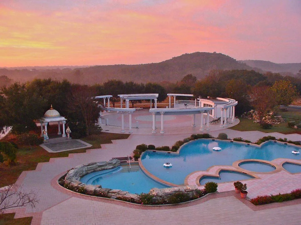 Spacious outdoor swimming pool area at sunset, with a kidney-shaped pool, smaller circular pools, and white colonnaded structures, surrounded by landscape with trees and hills in the background.