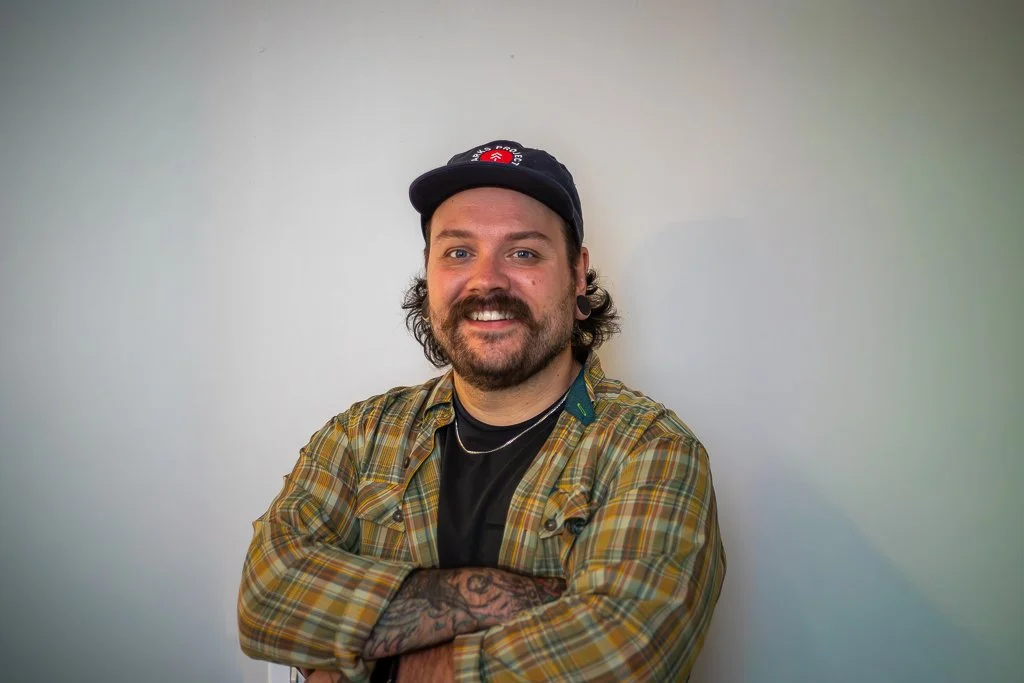 A man with a beard and tattoos wearing a plaid shirt and a baseball cap, smiling and crossing his arms, standing against a plain wall.
